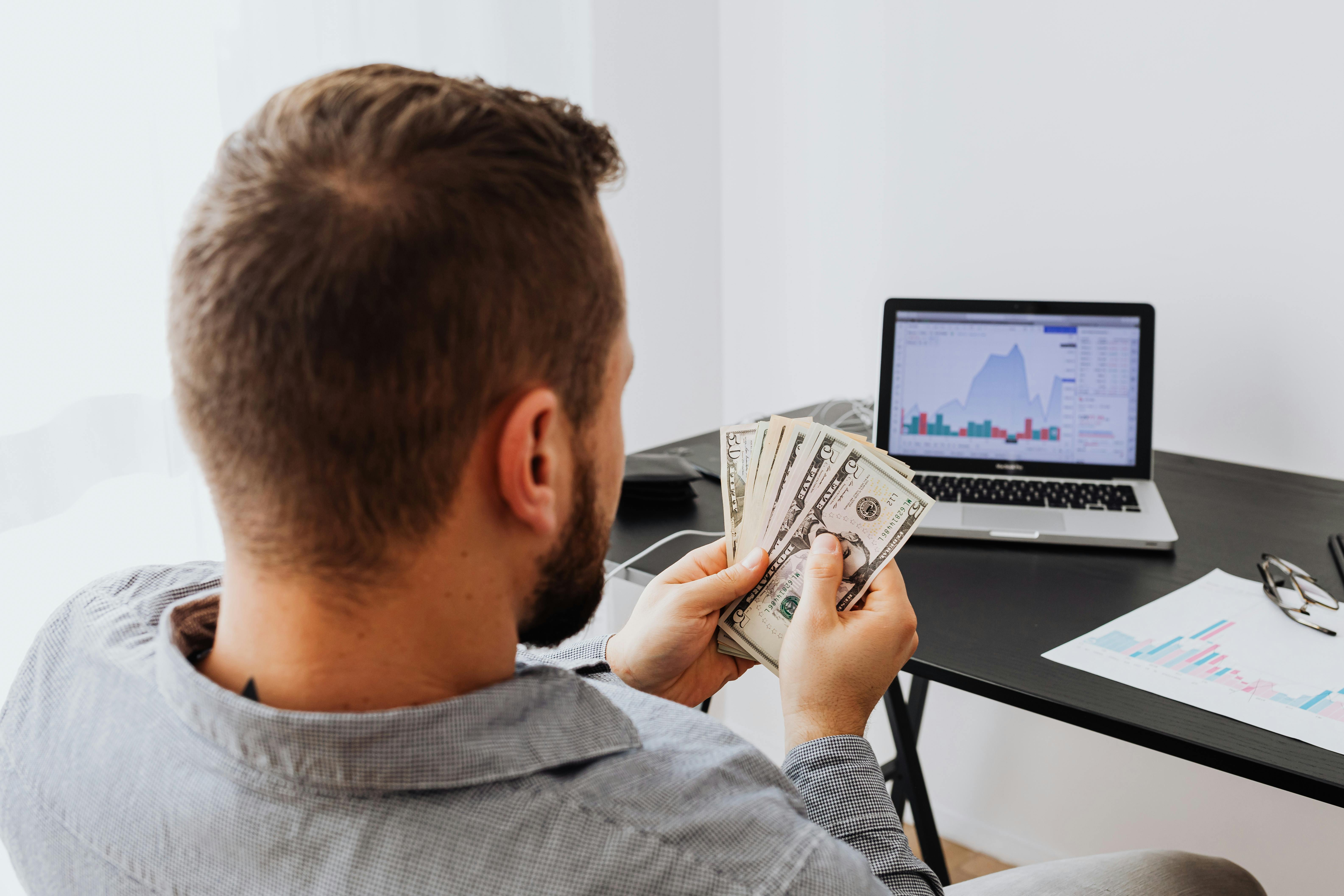 Ein Mann sitzt vor einem Laptop und zählt Geld | Quelle: Pexels