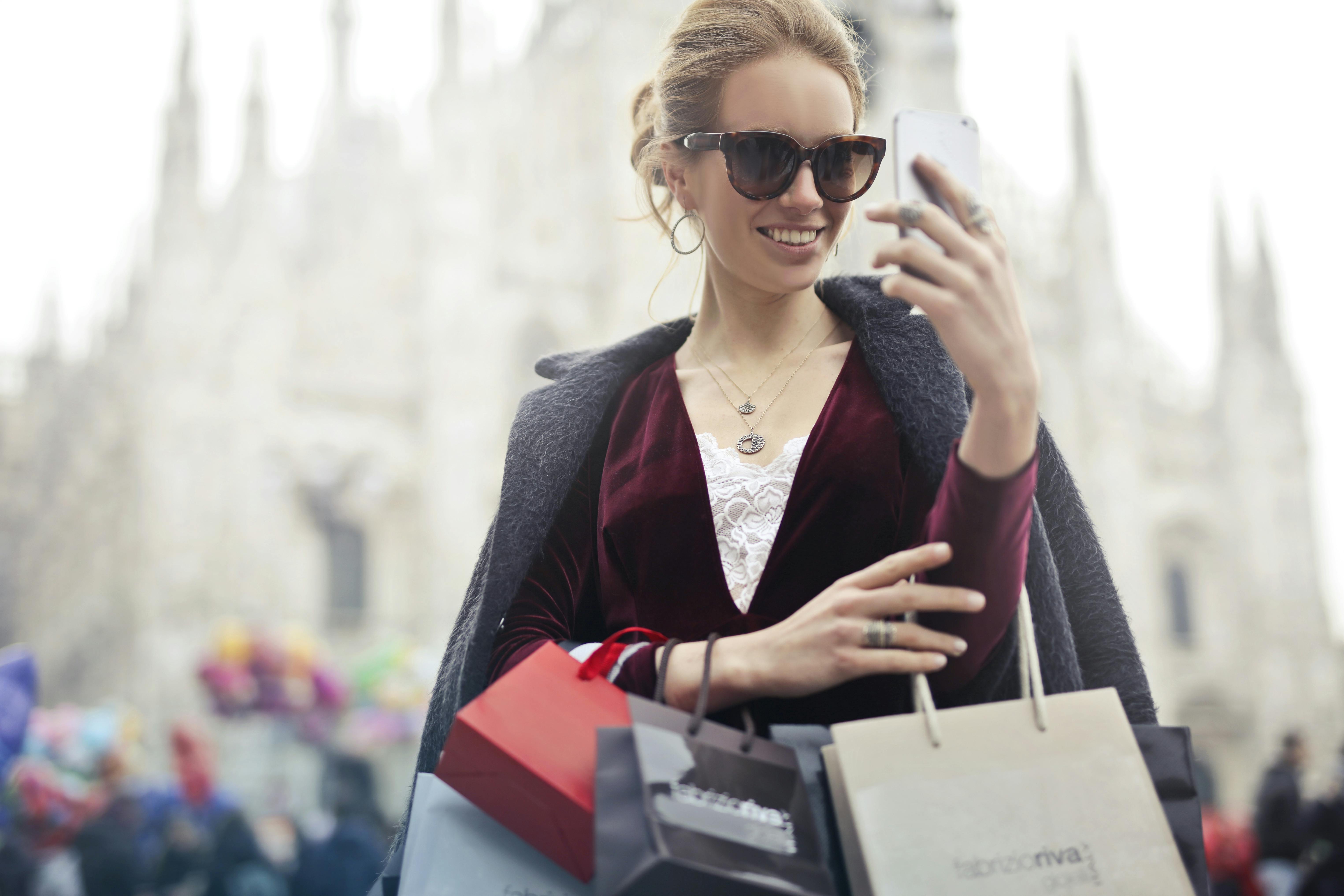 Eine wohlhabende Frau mit Sonnenbrille, die Einkaufstüten in der Hand hält und auf ihr Telefon schaut | Quelle: Pexels