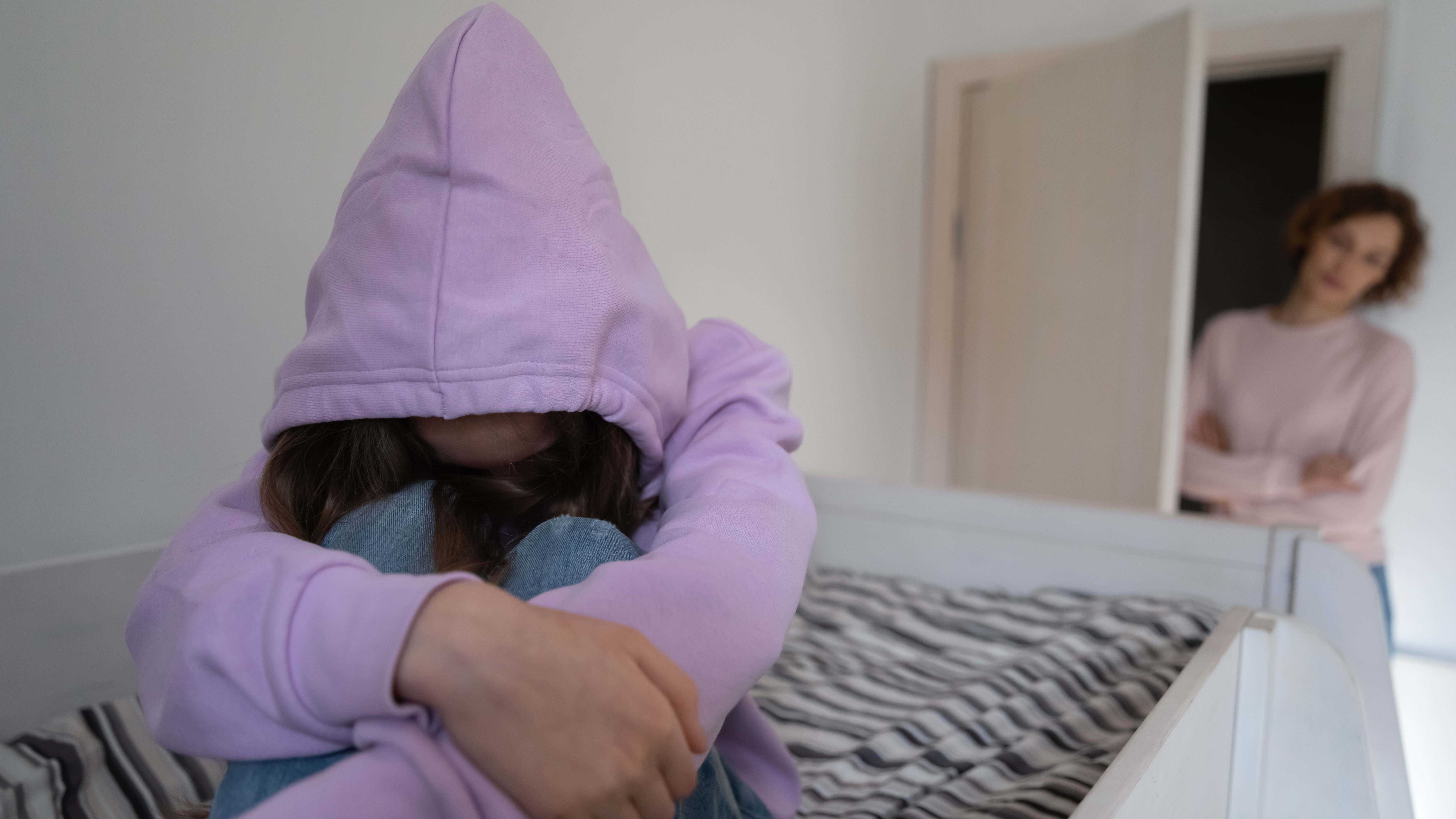 Ein junges Mädchen sitzt auf einem Bett und umarmt ihre Knie, während eine Frau von der Tür aus zusieht| Quelle: Shutterstock