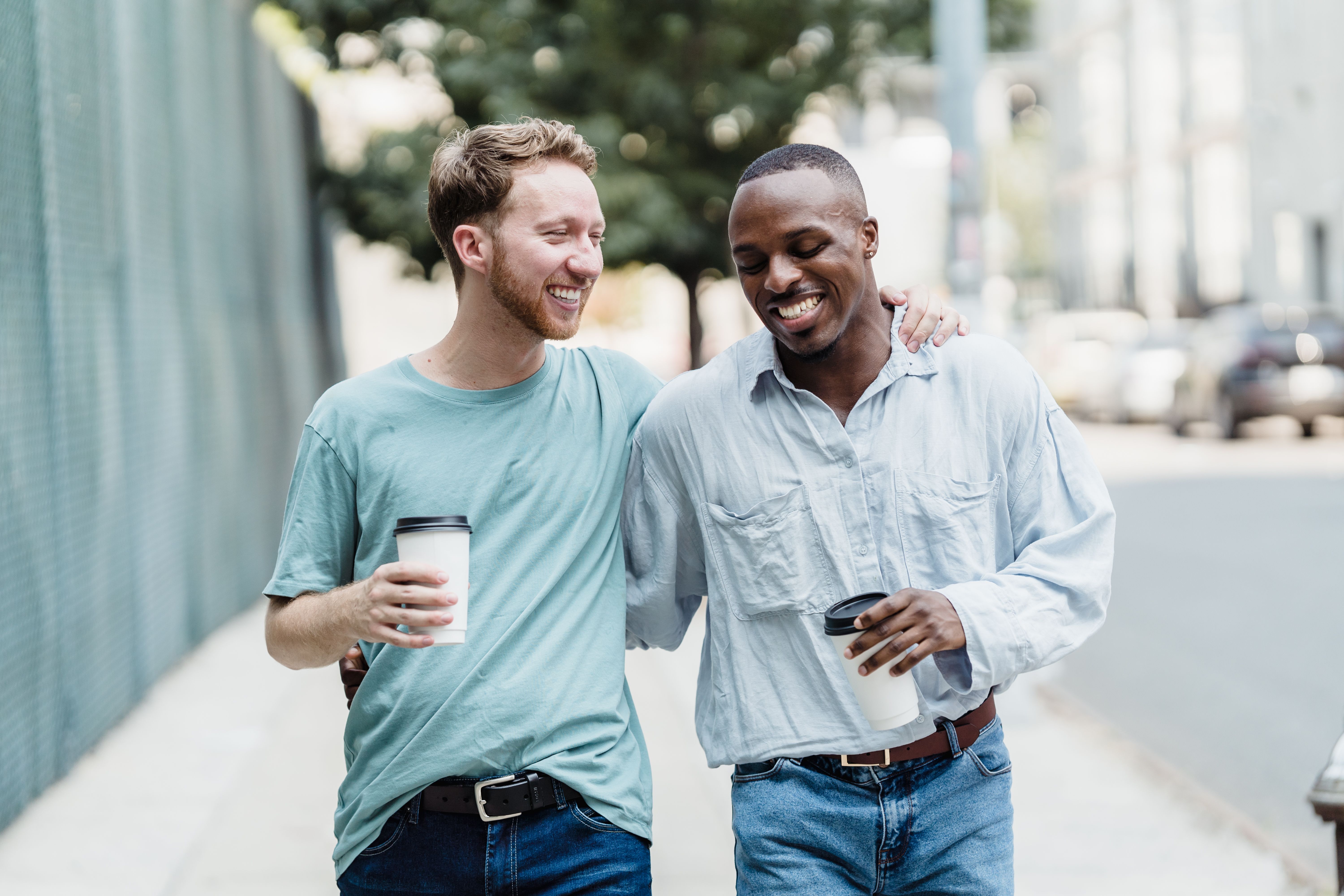 Zwei Freunde, die lachend durch eine Straße in der Stadt spazieren und gemeinsam im Freien einen Kaffee genießen | Quelle: Pexels