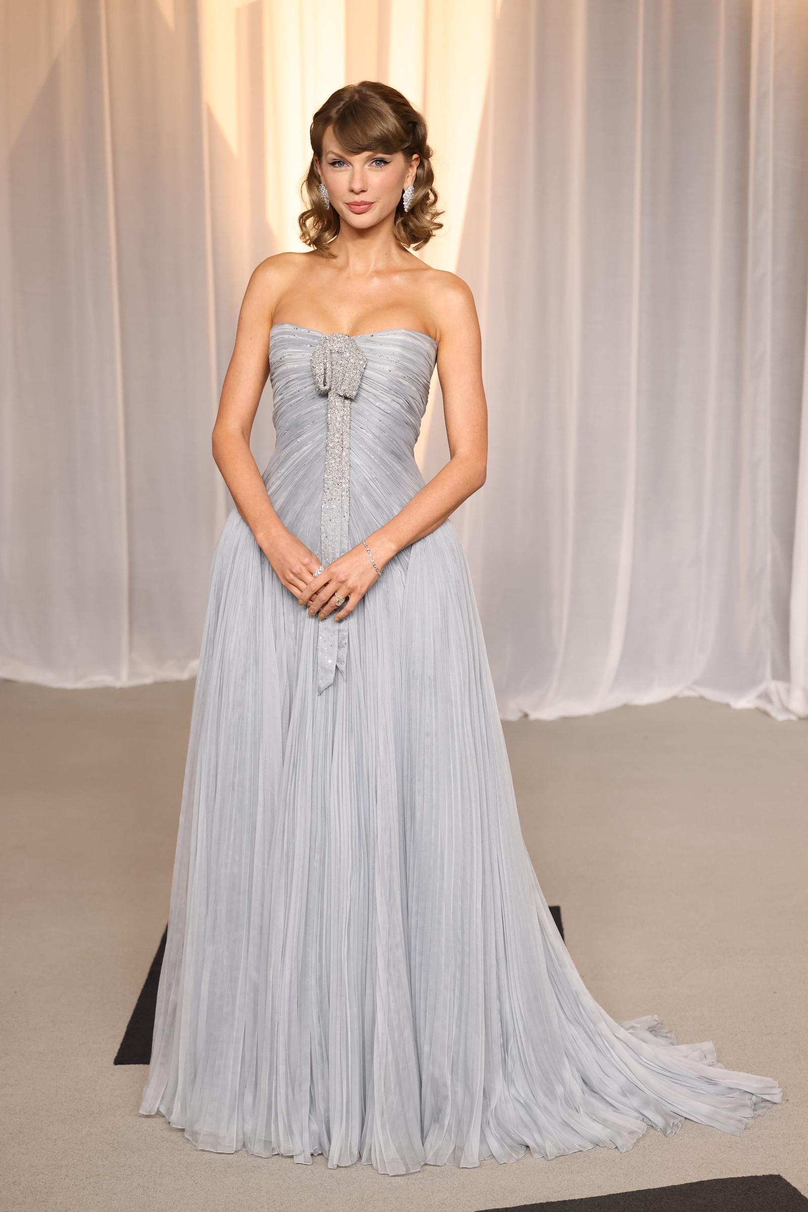 Taylor Swift posiert in einem trägerlosen, eisblauen Plissee-Kleid mit einer mit Kristallen besetzten Schleife und einer weit ausladenden Schleppe, dazu trägt sie funkelnde Ohrringe, ein Armband und Ringe.