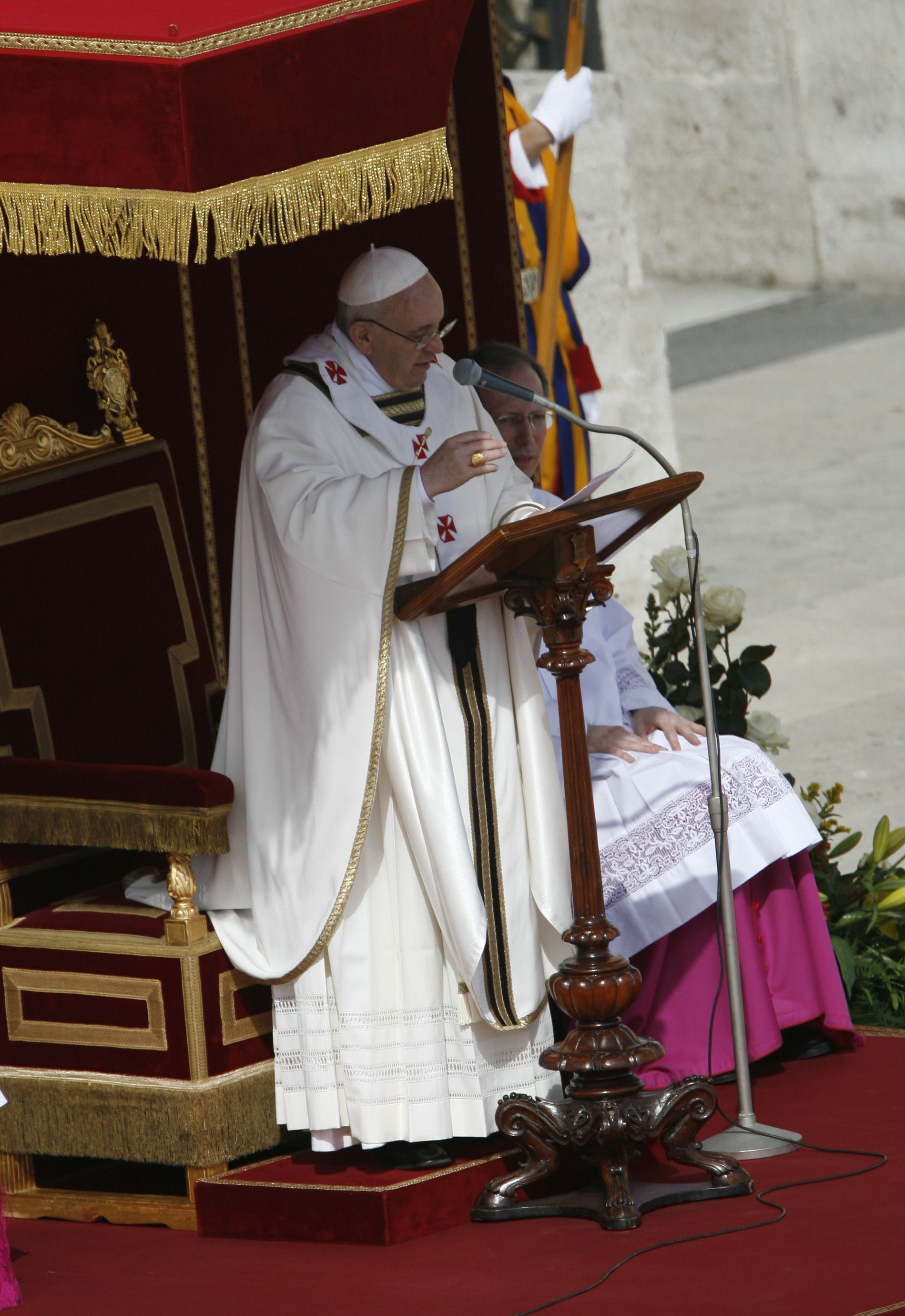 Papst Franziskus hält eine Predigt von einem geschnitzten Holzpult aus, in cremefarbene Gewänder mit roten Kreuzen gehüllt, neben einem thronähnlichen, mit Gold verzierten Stuhl stehend