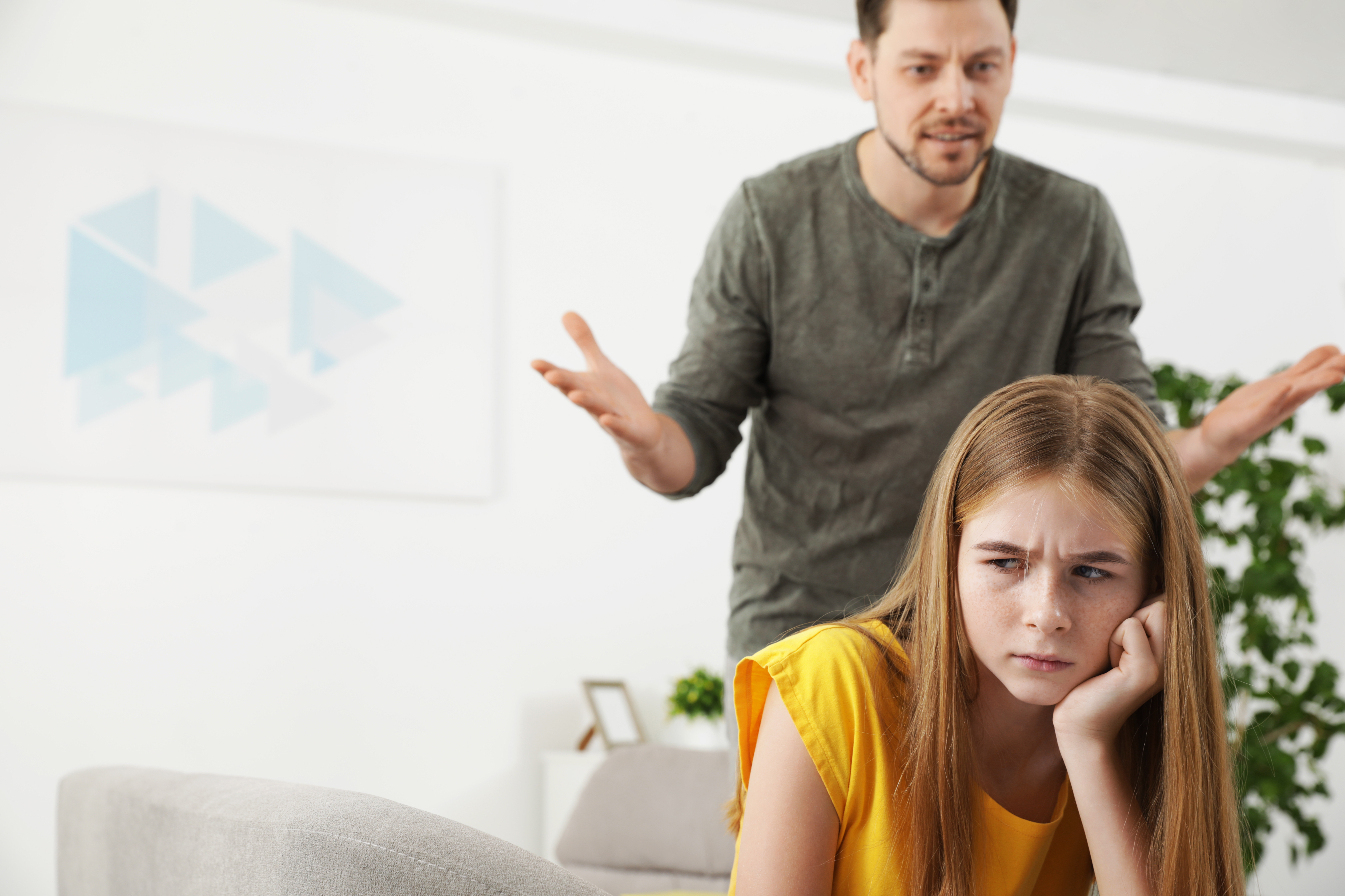 Ein wütender Mann steht hinter einem Mädchen, das auf einer Couch sitzt | Quelle: Shutterstock