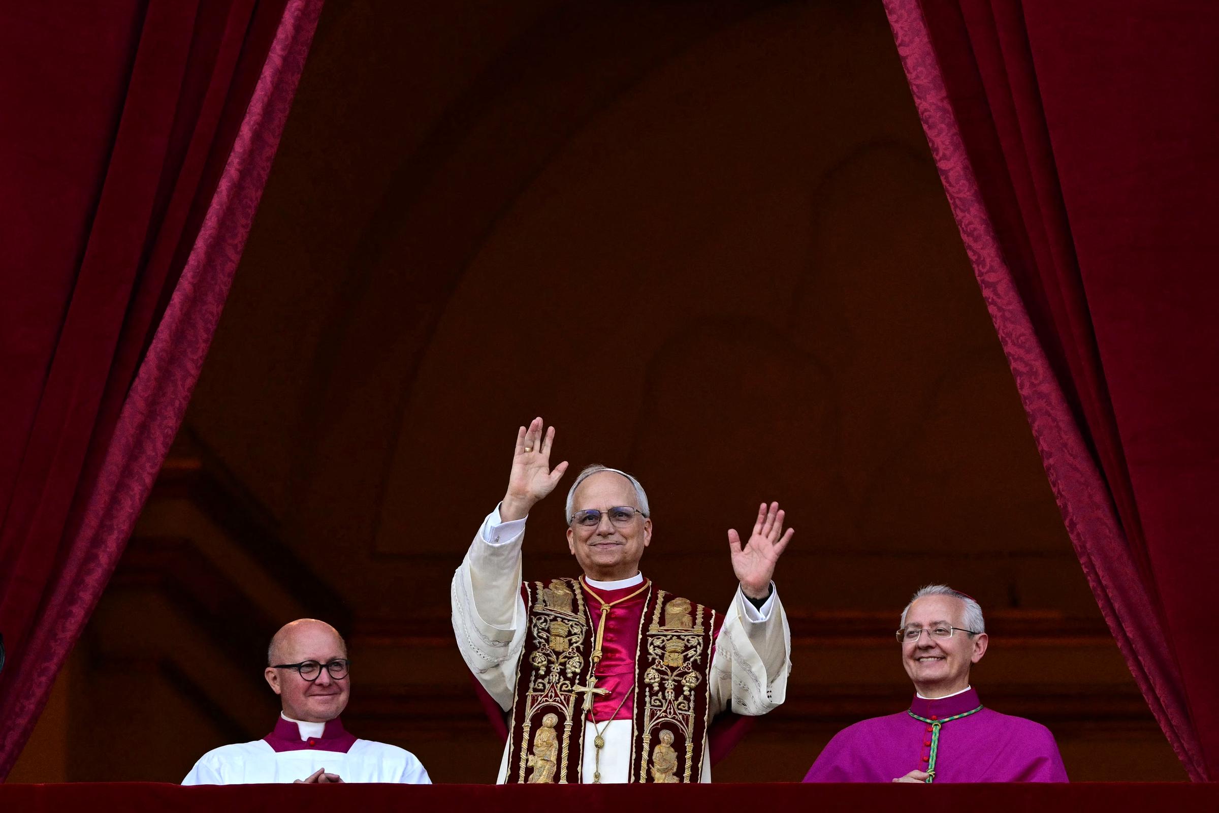 Papst Leo XIV. hebt zur Begrüßung beide Hände und trägt eine reich bestickte Stola über cremefarbenen Gewändern, als er auf den von tiefroten Samtvorhängen eingerahmten Balkon tritt