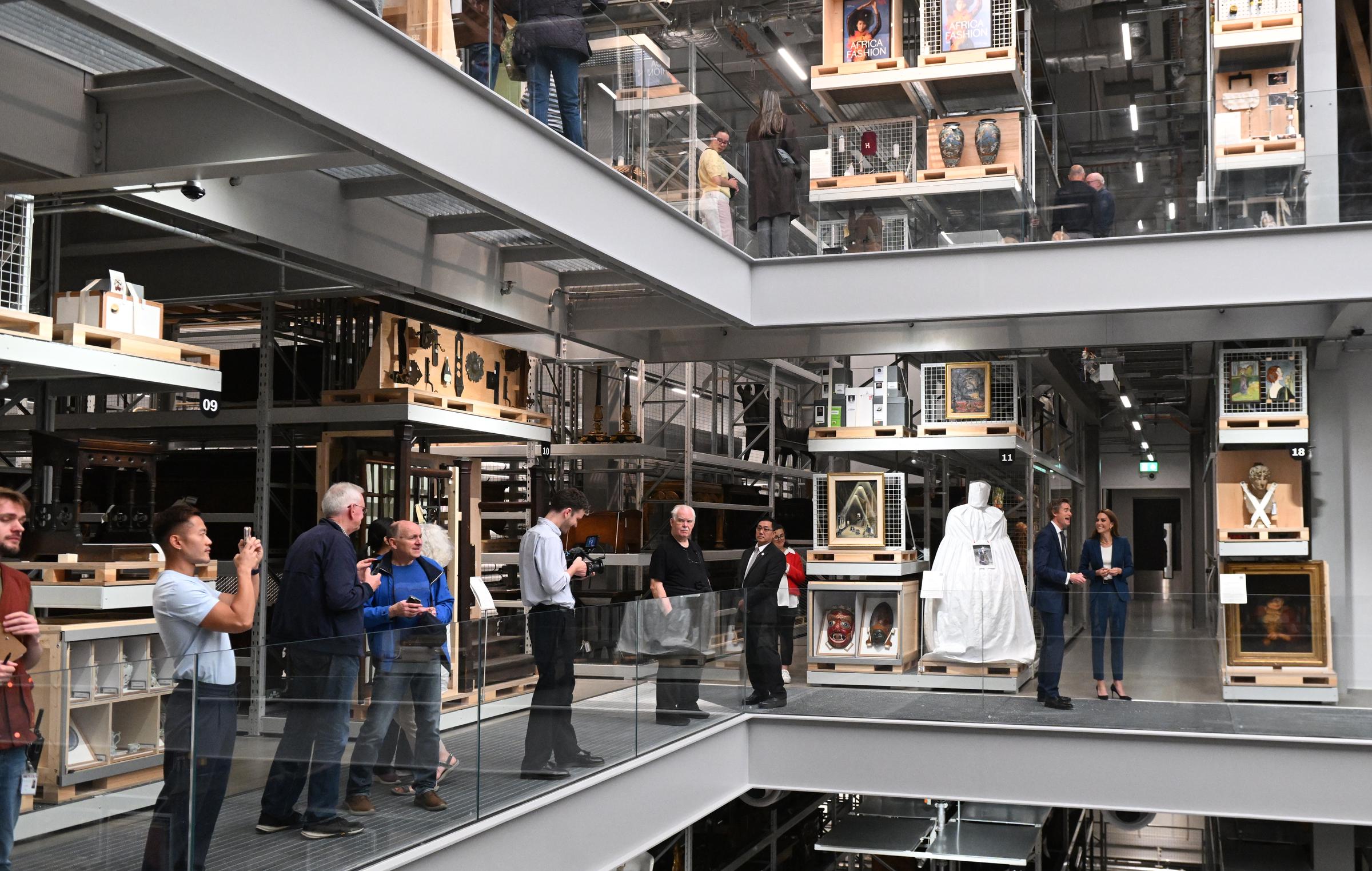 Inmitten einer geschäftigen Kulisse von Besuchern und Mitarbeitern geht Prinzessin Catherine mit Tristram Hunt durch das V&A East Storehouse. In ihrem königsblauen Anzug wirkt sie gelassen und aufmerksam, während sie durch die beeindruckenden, mehrstöckigen Galerieräume geht.