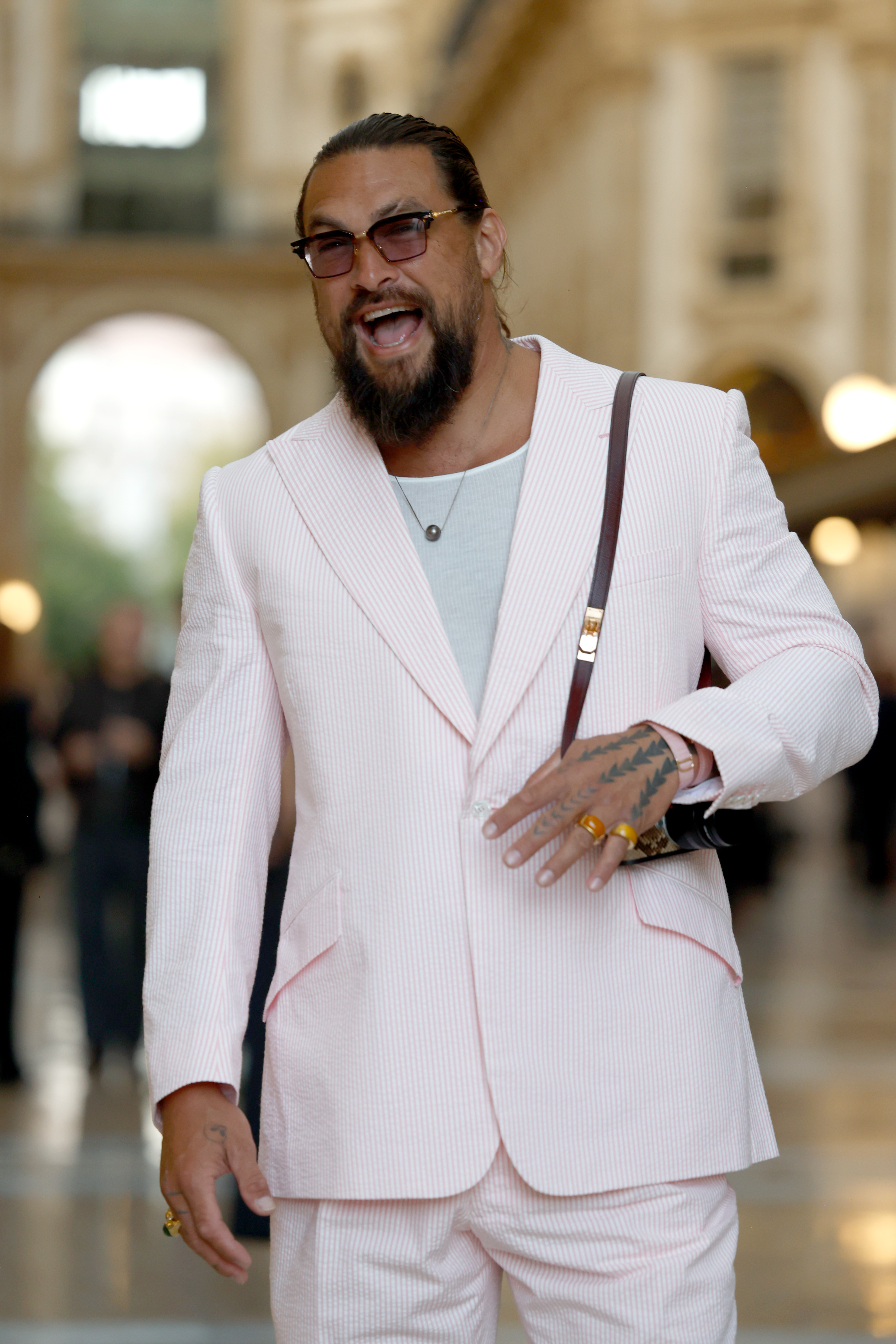 Momoa lacht, während er im Saal posiert, und gibt dabei einen genaueren Blick auf seine getönten Brillen, seinen Schmuck und die feinen rosa Streifen auf seinem Anzug frei. | Quelle: Getty Images