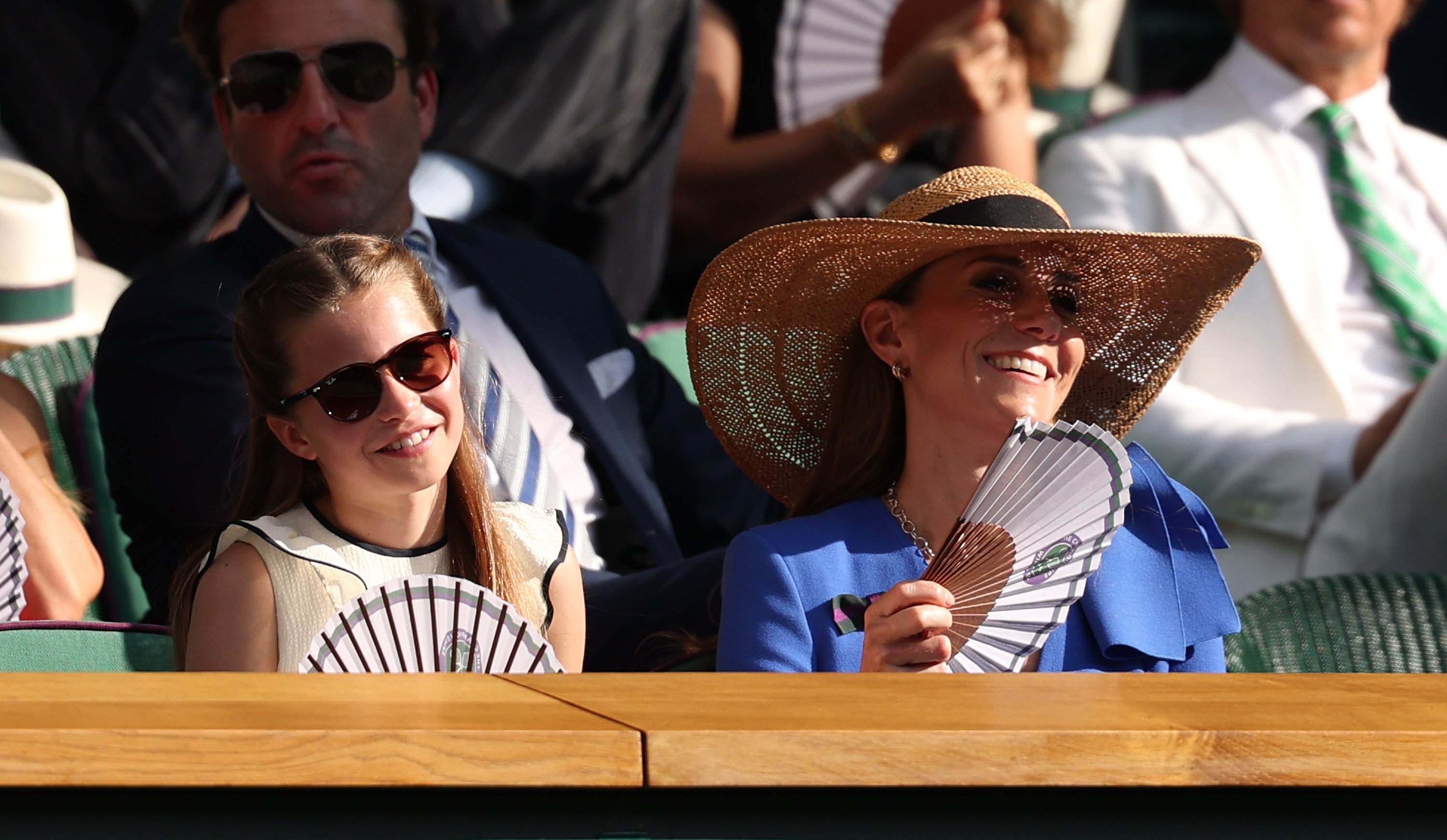 Prinzessin Charlotte von Wales und Catherine, Prinzessin von Wales, lächeln, während sie sich am vierzehnten Tag der Wimbledon-Meisterschaften am 13. Juli 2025 im All England Lawn Tennis and Croquet Club in London, England, mit Papierfächern Abkühlung verschaffen. Charlotte verfolgt das Finale im Herreneinzel mit einer dunklen Sonnenbrille in Schildpattoptik und einem cremefarbenen, ärmellosen Oberteil mit Rüschen. Sie sitzt neben Catherine, die in einem leuchtend blauen Kleid, einer auffälligen Sonnenbrille und einem breitkrempigen Strohhut sommerlich aussieht. | Quelle: Getty Images