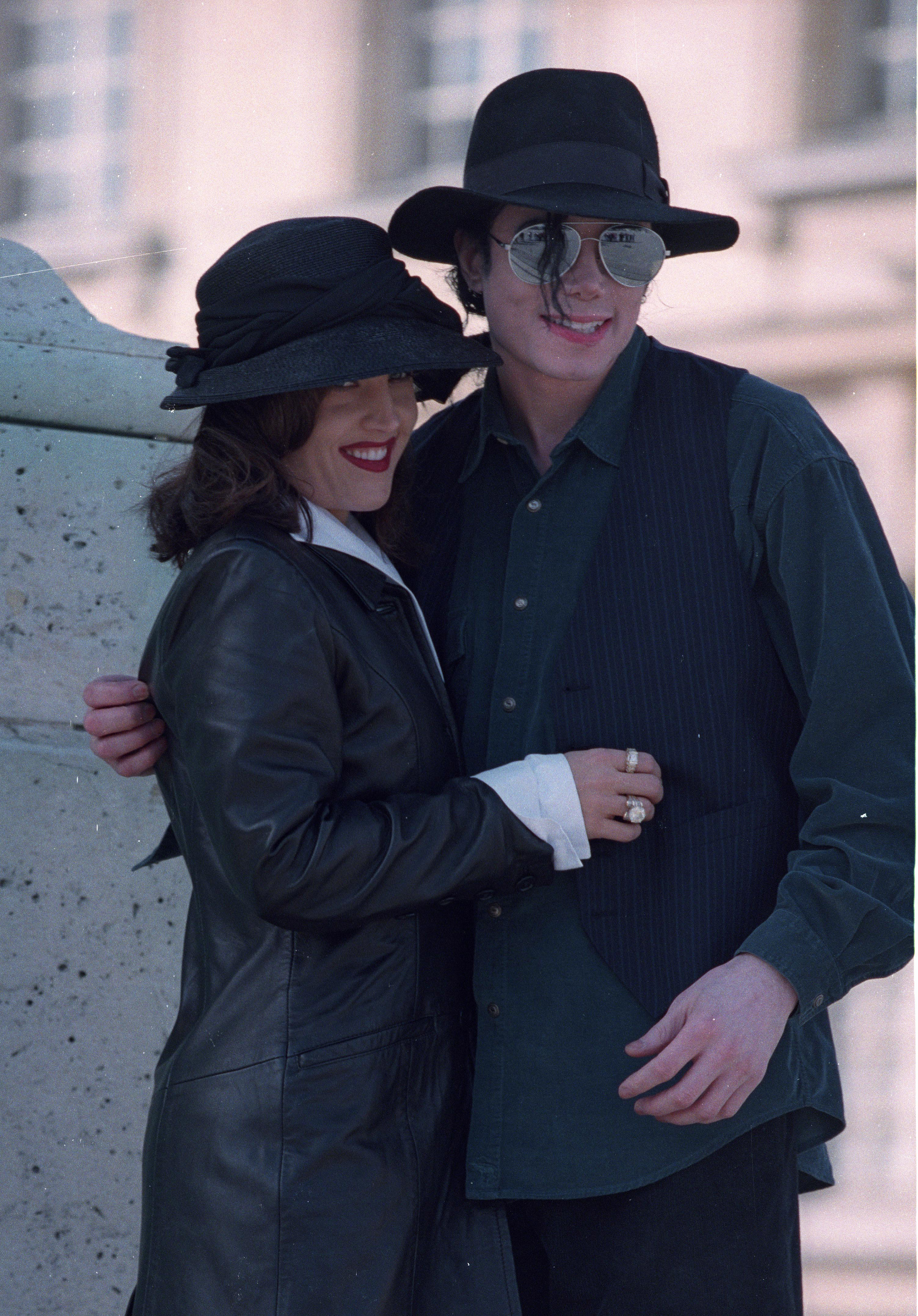 Lisa Marie Presley umarmt ihren Ehemann Michael Jackson bei einem Besuch in Versailles, Frankreich, im Jahr 1995. Sie trägt einen eleganten schwarzen Ledermantel und einen schwarzen Hut mit breiter Krempe, während Jackson mit einer Pilotenbrille, einem schwarzen Fedora und einem dunklen Hemd, das unter einer Nadelstreifenweste getragen wird, lächelt. | Quelle: Getty Images