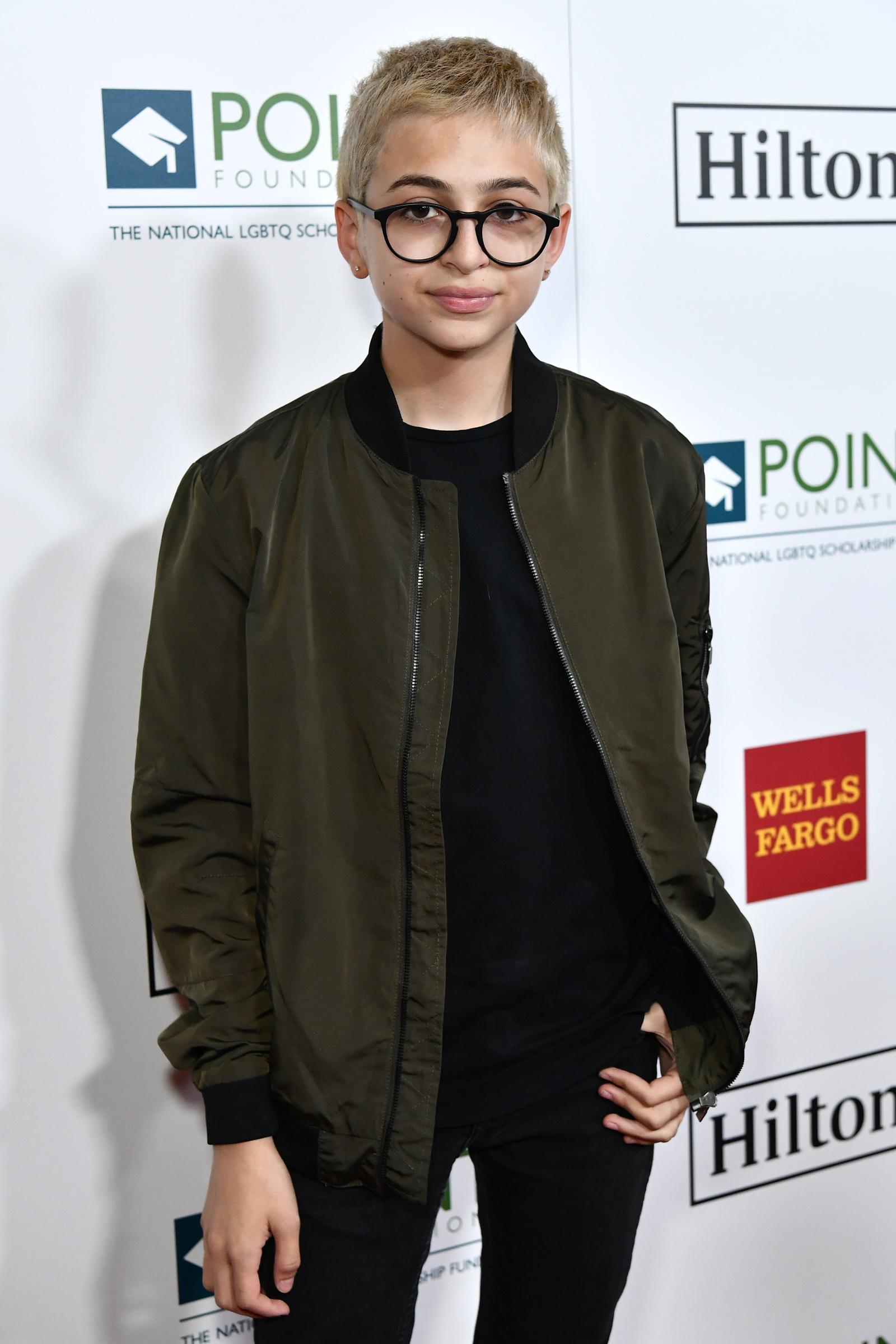 J.J. Totah besucht die Point Honors Gala in Los Angeles, Kalifornien, und trägt eine dunkle Bomberjacke über einem schwarzen Hemd, einer schwarzen Hose und einer runden Brille. Totahs kurzes, helles Haar rundet den Look ab. | Quelle: Getty Images