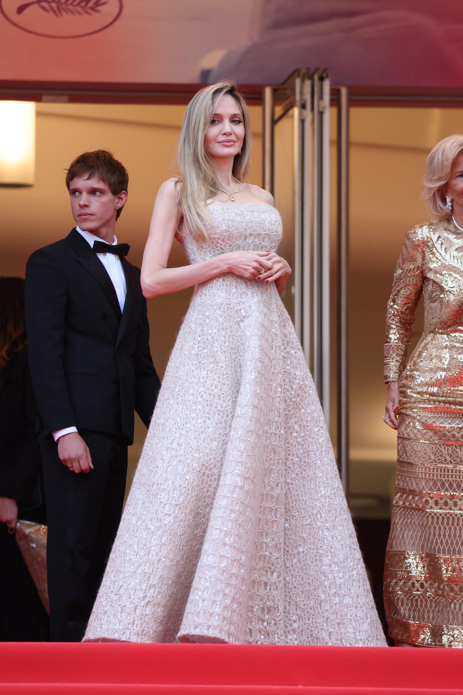Angelina Jolie steht ganz oben auf der Treppe des Palais des Festivals, die Hände sanft vor ihrem aufwändig gearbeiteten Kleid von Brunello Cucinelli verschränkt und strahlt zeitlose Gelassenheit vor einem Hintergrund aus Rot und Gold aus.
