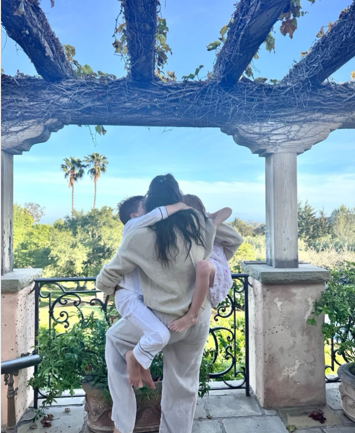 Meghan Markle steht unter einer mit Weinreben bewachsenen Pergola und umarmt Prinz Archie und Prinzessin Lilibet in einer fröhlichen Familienumarmung. | Quelle: Instagram/meghan