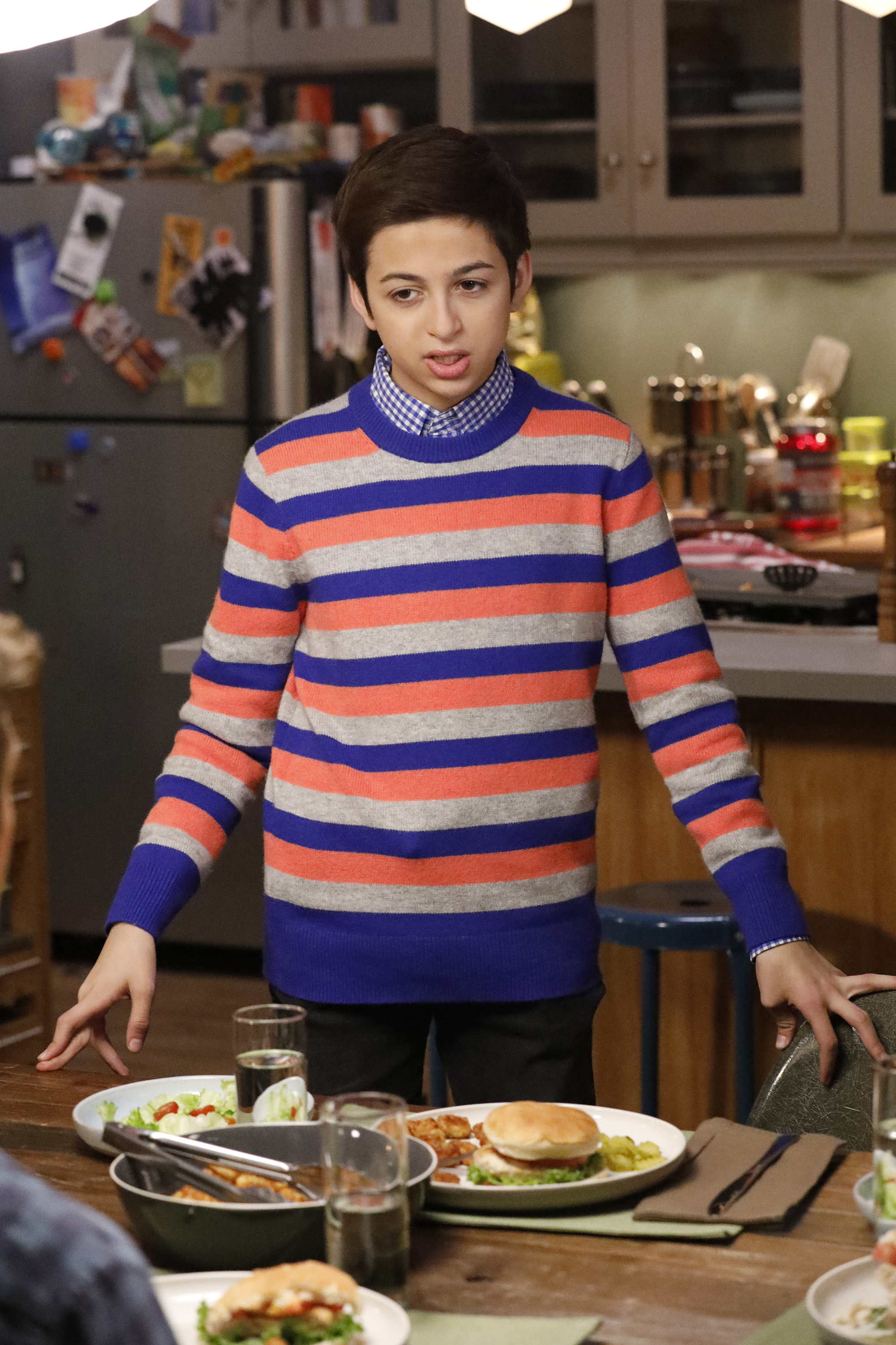 J.J. Totah spielt Michael in der Folge von „Champions“ „Vincemas“, trägt einen bunten gestreiften Pullover über einem blau karierten Hemd und steht neben einem Esstisch in einer Küche. | Quelle: Getty Images