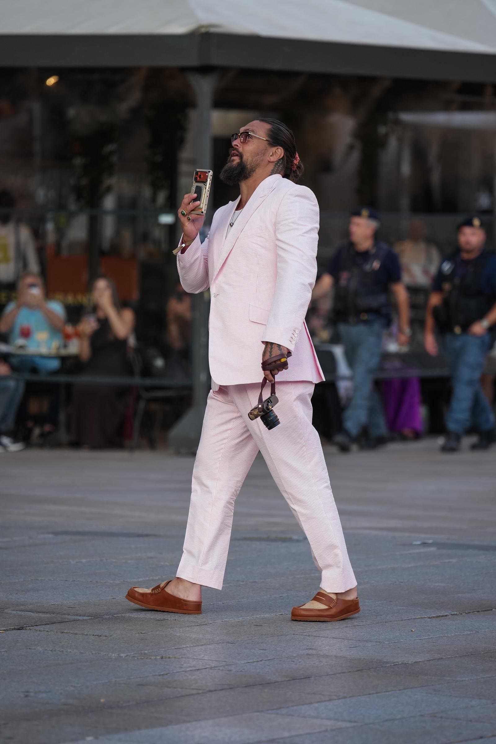 Momoa geht nach draußen und hält dabei sein Handy hoch, wodurch die lockeren Schnitte des Anzugs und seine braunen Slipper gut zur Geltung kommen. | Quelle: Getty Images