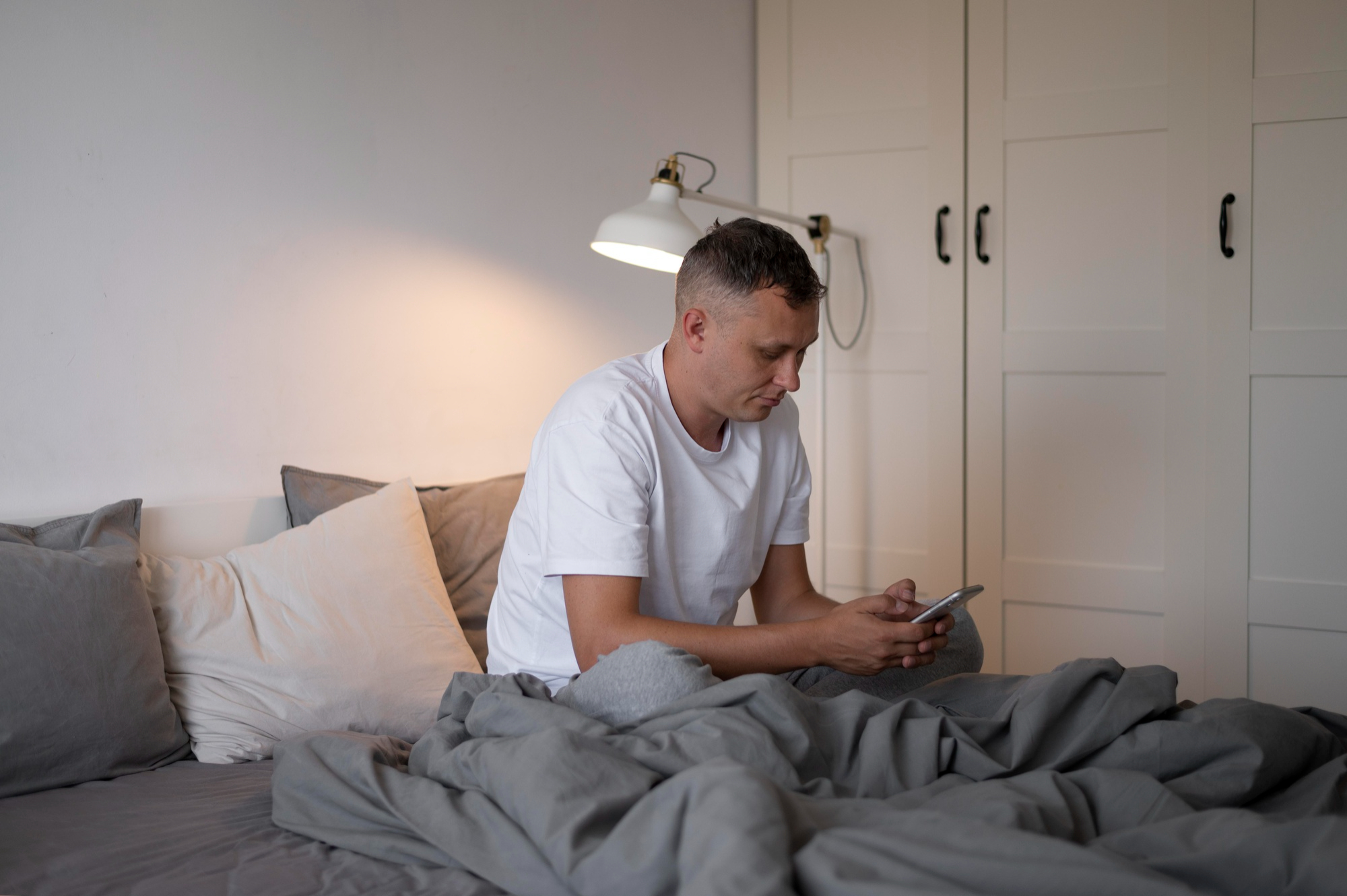 Ein Mann schaut auf sein Telefon, während er mit einem Laken zugedeckt im Bett liegt | Quelle: Freepik