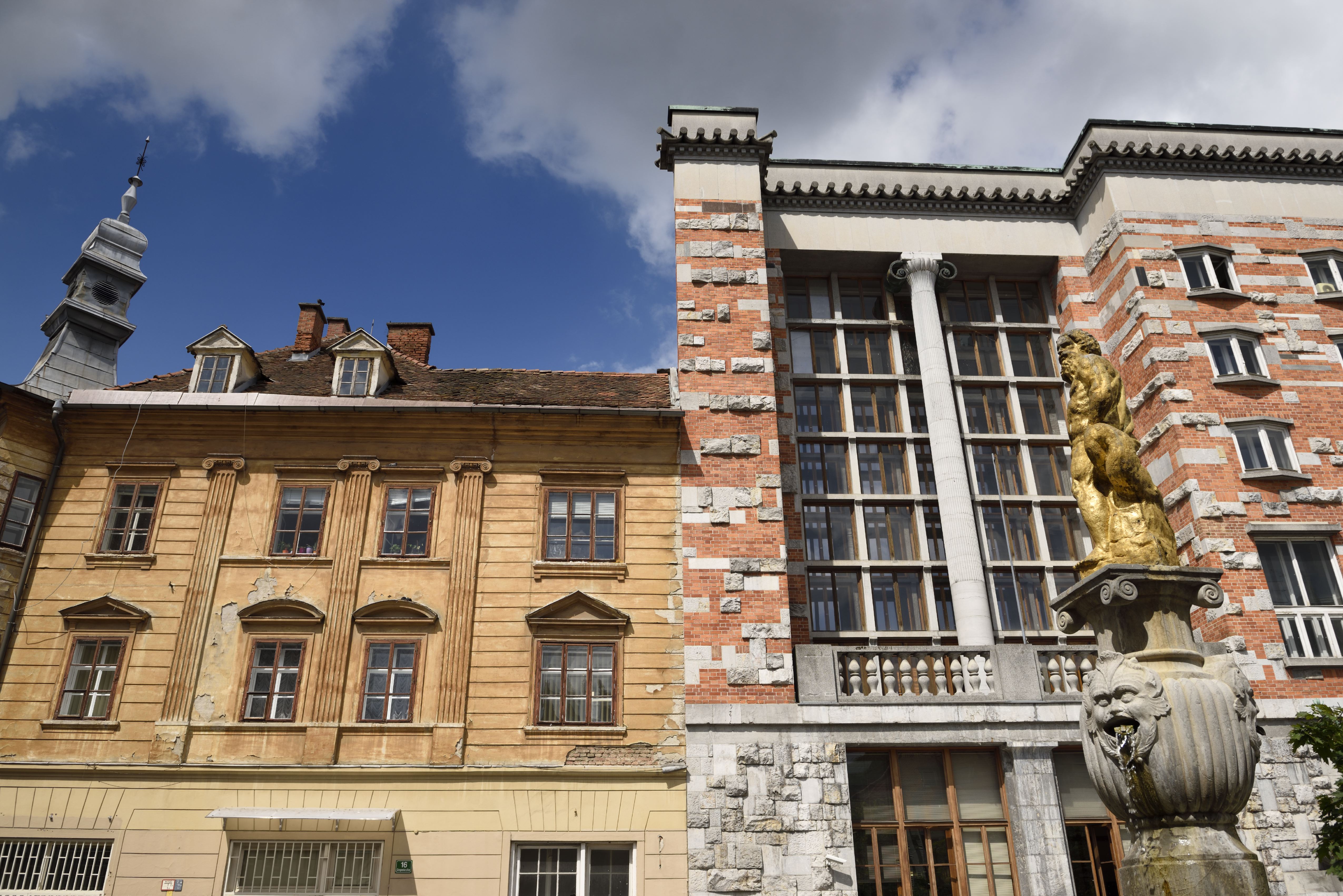 Die goldene Neptun-Statue auf dem Brunnen von Janez Kumersteiner aus dem Jahr 1675 steht im Vordergrund vor der markanten Fassade aus rotem Backstein und Stein der National- und Universitätsbibliothek Sloweniens in Ljubljana. Das vom berühmten Architekten Jože Plečnik entworfene, viel gepriesene Bibliotheksgebäude erhebt sich unter strahlend blauem Himmel neben dem benachbarten klassischen gelben Stadthaus und der Vžigalica-Galerie. | Quelle: Getty Images