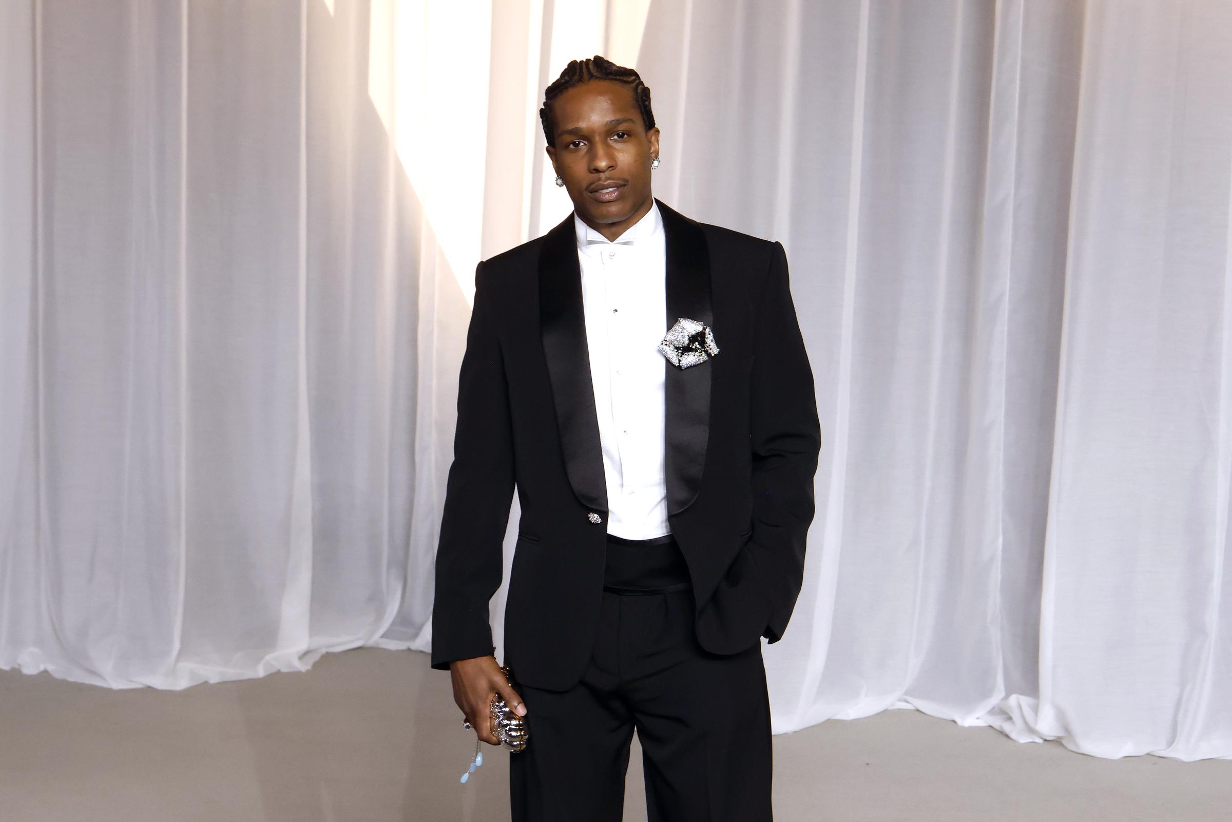 A$AP Rocky verkörpert moderne Eleganz im Black-Tie-Stil in einem schnittigen Smoking mit breiten Satin-Schalrevers und einem hoch sitzenden Kummerbund. Statt einer traditionellen Fliege trägt der Rapper ein minimalistisches weißes Hemd mit Flügelkragen und wertet das klassische Ensemble mit einer kunstvollen Blumenbrosche am Revers, gepflegten Cornrows und einer funkelnden, ausgefallenen Abend-Clutch auf.