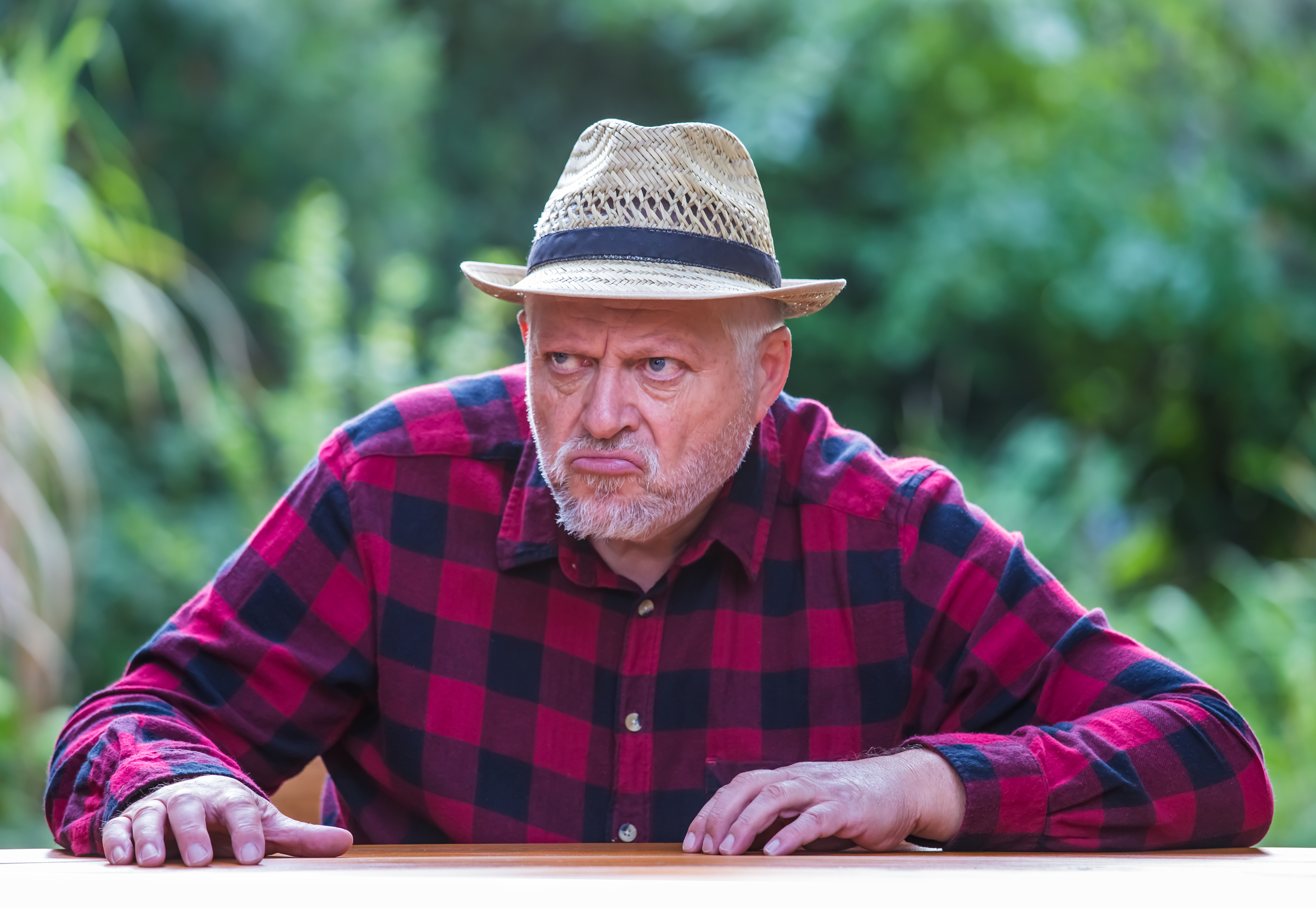 Ein älterer Mann mit Hut sitzt mit schlechter Laune an einem Tisch im Garten. | Quelle: Shutterstock