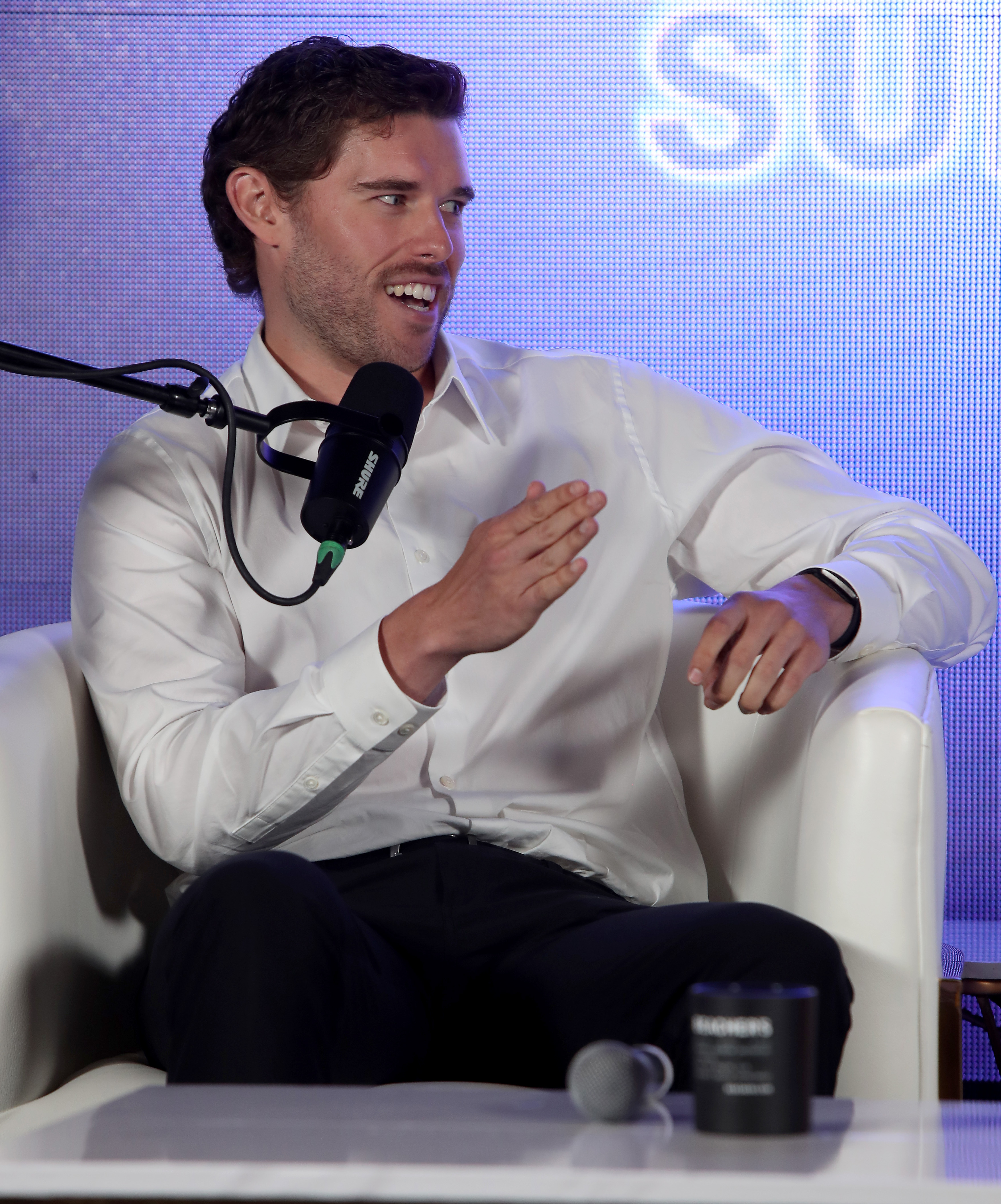 Christopher Schwarzenegger ist mitten in einer Diskussion während einer Podiumsdiskussion auf dem Beacher Vitality Happy & Healthy Summit zu sehen. Er trägt ein maßgeschneidertes weißes Hemd und eine maßgeschneiderte schwarze Hose. Sein auffallend schlanker Körper spiegelt die Ergebnisse seiner jahrelangen gesundheitlichen Transformation wider.