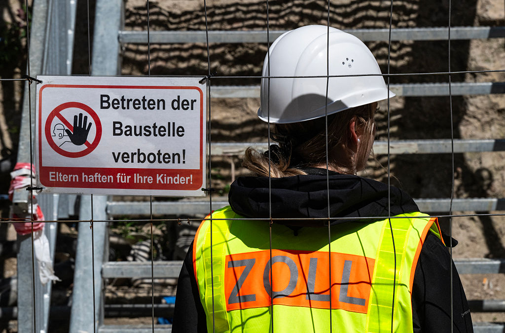 Ein Beamter des deutschen Zollamts, der einen Helm trägt, überwacht eine Baustelle, während seine Kollegen nach illegalen Arbeitskräften suchen; auf einem Schild am Zaun steht „Das Betreten der Baustelle ist verboten“ – Berlin, 6. August 2025 | Quelle: Getty Images