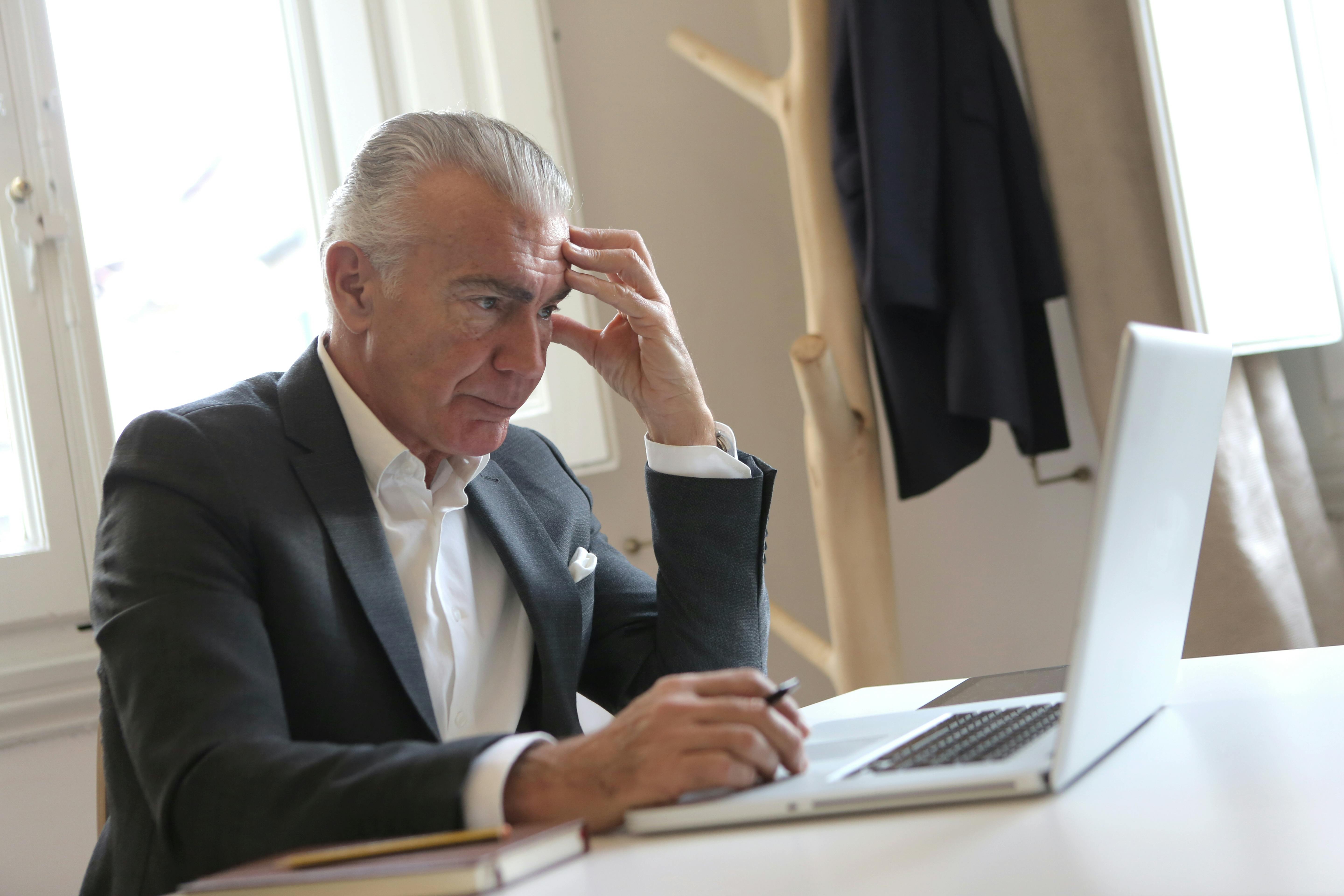 Ein gestresster älterer Mann sitzt mit der Hand an der Schläfe, während er einen Stift hält und auf seinen Laptop schaut | Quelle: Pexels