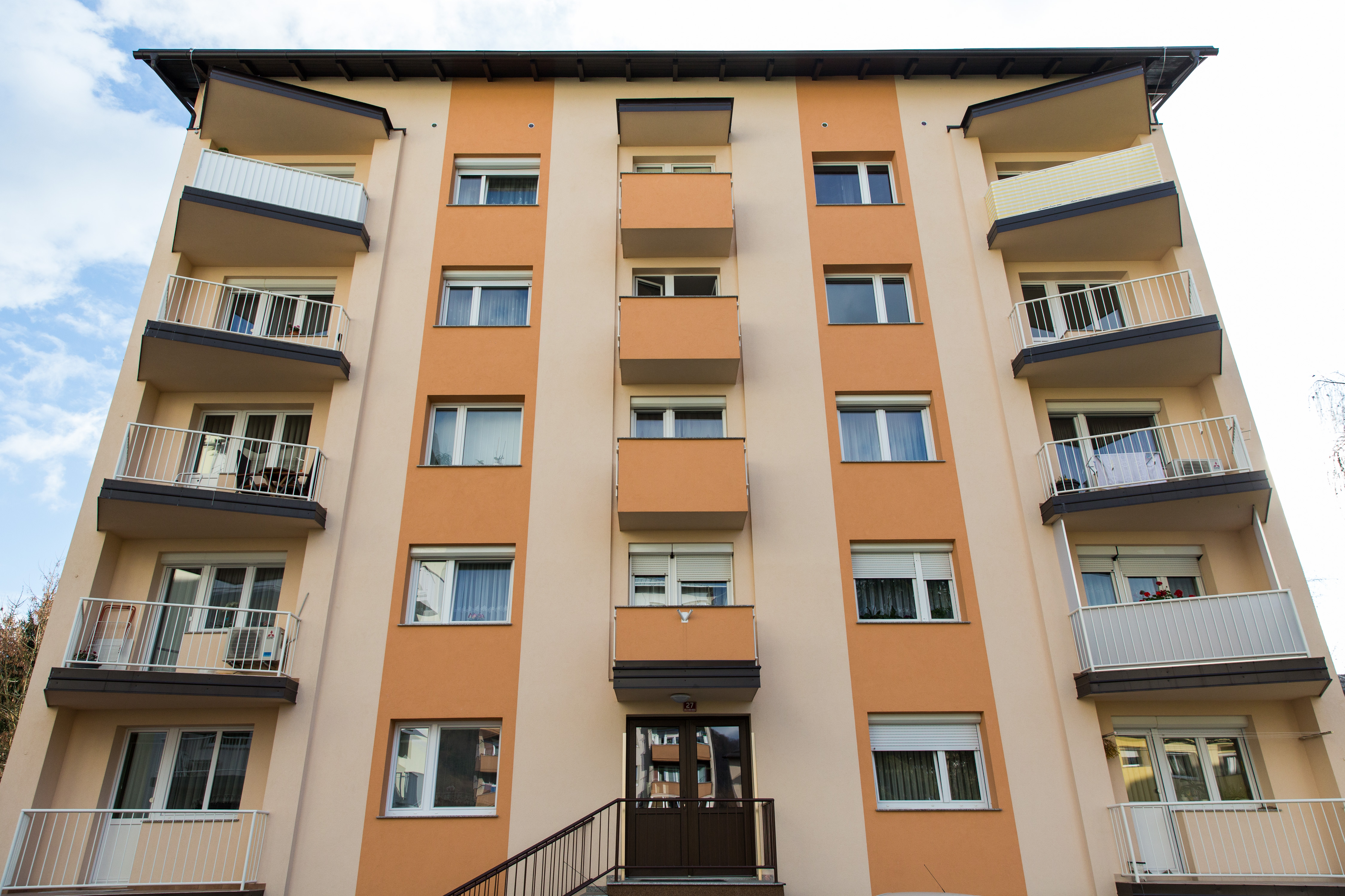 Eine direkte Aufnahme aus niedrigem Blickwinkel zeigt die symmetrische Fassade des Wohnkomplexes in Sevnica, Slowenien, in dem Melania Trump aufgewachsen ist. Das Foto wurde am 28. November 2016 aufgenommen. Die Fassade besticht durch warme orangefarbene vertikale Akzente, ordentlich angeordnete Balkongeländer und eine zentrale Eingangstreppe, die in das ruhige Wohngebäude führt. | Quelle: Getty Images