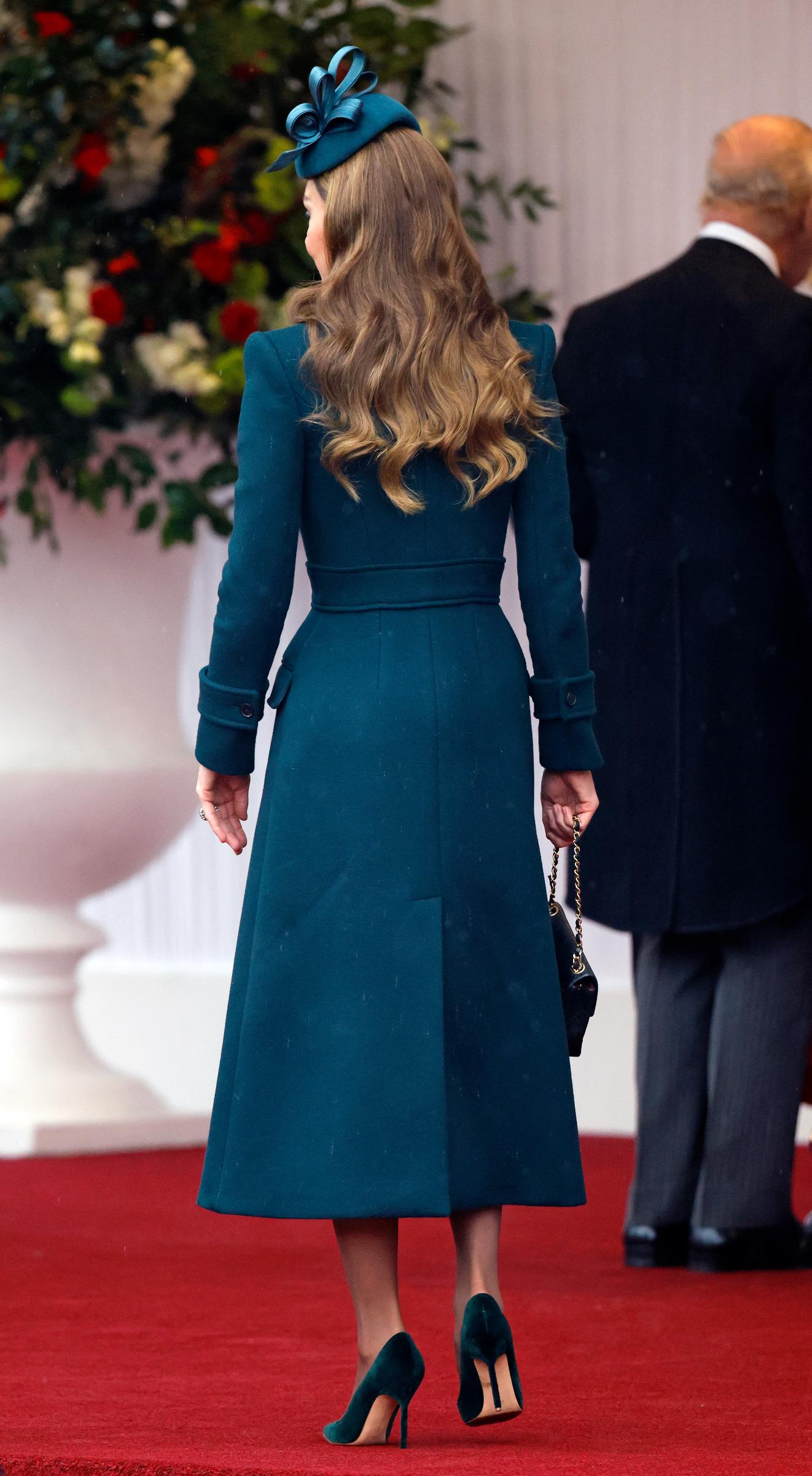 Ein Blick auf Catherine, Prinzessin von Wales, von hinten unterstreicht die klaren Linien ihres maßgeschneiderten Midi-Mantelkleids, als sie am 20. Oktober 2026 in Windsor über den roten Teppich schreitet. Die Rückseite des tiefgrünen Outfits zeigt einen markanten Taillengürtel und eine mittige Schlitzfalte, ergänzt durch sanfte brünette Locken, die unter einem Fascinator von Juliette Millinery herabfallen, samtige Stiletto-Pumps und eine gesteppte Chanel-Handtasche, die sie am Kettenriemen trägt. | Quelle: Getty Images