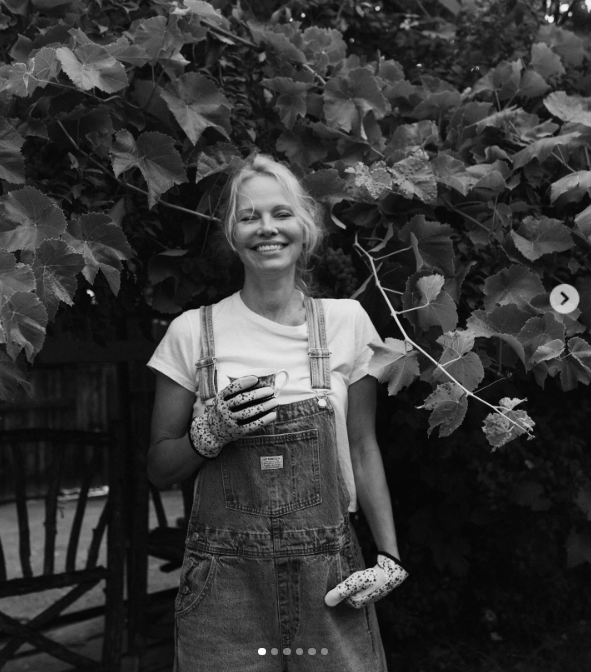 Auf einem strahlenden Schwarz-Weiß-Porträt zeigt Pamela Anderson vor einer dichten Wand aus Weinblättern im Arcady ein strahlendes, ungeschminktes Lächeln. Sie trägt eine Jeanslatzhose, Gartenhandschuhe und hält eine kleine Teetasse in der Hand. Dabei strahlt sie pure Freude aus, während sie über „Pamela’s Cooking with Love“ spricht und die Show auf Vancouver Island als ein echtes Traumprojekt bezeichnet, das ihren blühenden Garten mit internationalem kulinarischem Talent verbindet. | Quelle: Instagram/pamelaanderson