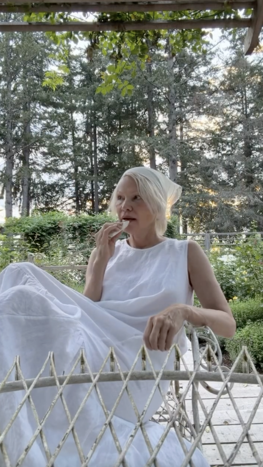 Pamela Anderson faulenzt gemütlich in einem Vintage-Gartenstuhl in Arcady, gekleidet in ein gestuftes weißes, ärmelloses Kleid und mit einem cremefarbenen Kopftuch, das sie sich um ihr blondes Haar gebunden hat. Während sie in die Ferne blickt und ganz entspannt einen Happen isst, verkörpert sie das ruhige, geerdete Leben auf dem Anwesen, versteckt zwischen den hohen Kiefern von British Columbia, und teilt diesen Moment mit dem Kommentar: „Lebe romantisch, wo immer du bist… 🤍🌷“. | Quelle: Instagram/pamelaanderson