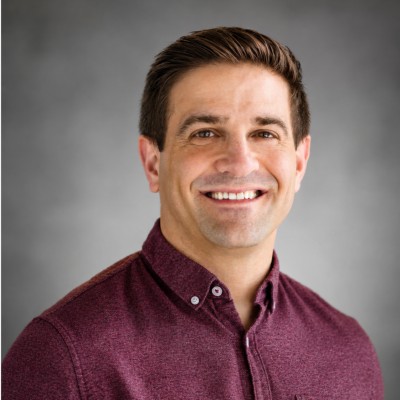 Tyler Zacharia lächelt auf einem Porträtfoto vor einem neutralen grauen Studiohintergrund. Er trägt ein zugeknöpftes, meliertes, kastanienbraunes Hemd mit Kragen und hat sein braunes Haar ordentlich mit Seitenscheitel frisiert. | Quelle: LinkedIn/Tyler Zacharia