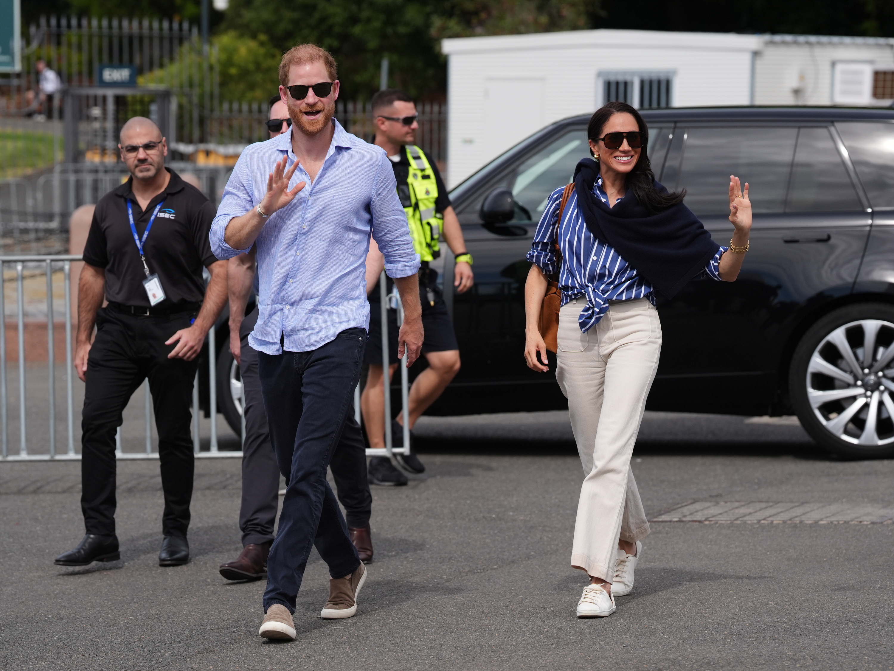 Meghan, Herzogin von Sussex, winkt gemeinsam mit Prinz Harry den Gratulanten zu, als sie am 17. April 2026 in Sydney, Australien, an den Man O’War Steps ankommen, vor einer Segelveranstaltung mit Mitgliedern von Invictus Australia. Sie trägt ein blau-weiß gestreiftes Hemd, das an der Taille gebunden ist, dazu eine cremefarbene Hose mit weitem Bein, eine Sonnenbrille, einen dunklen Strickpullover, der über ihre Schultern drapiert ist, und weiße Low-Top-Sneaker. | Quelle: Getty Images
