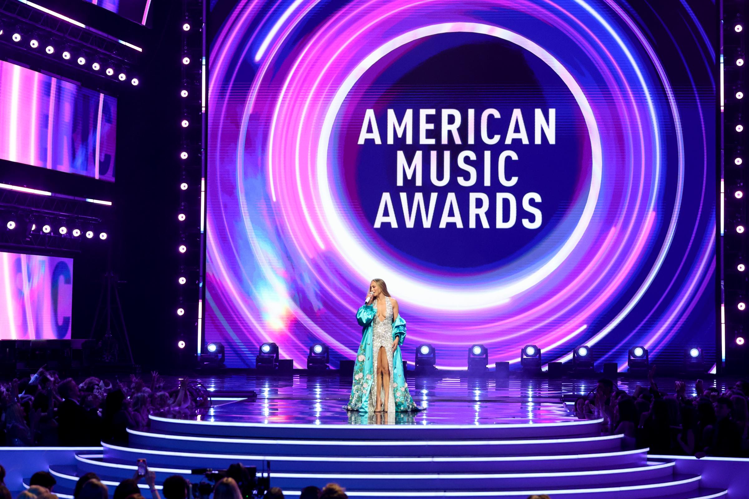 In einer Totalen der schillernden Bühne steht die 55-jährige Sängerin im Rampenlicht der American Music Awards, umrahmt von wirbelnden Neonlichtern, während das Publikum zuschaut.