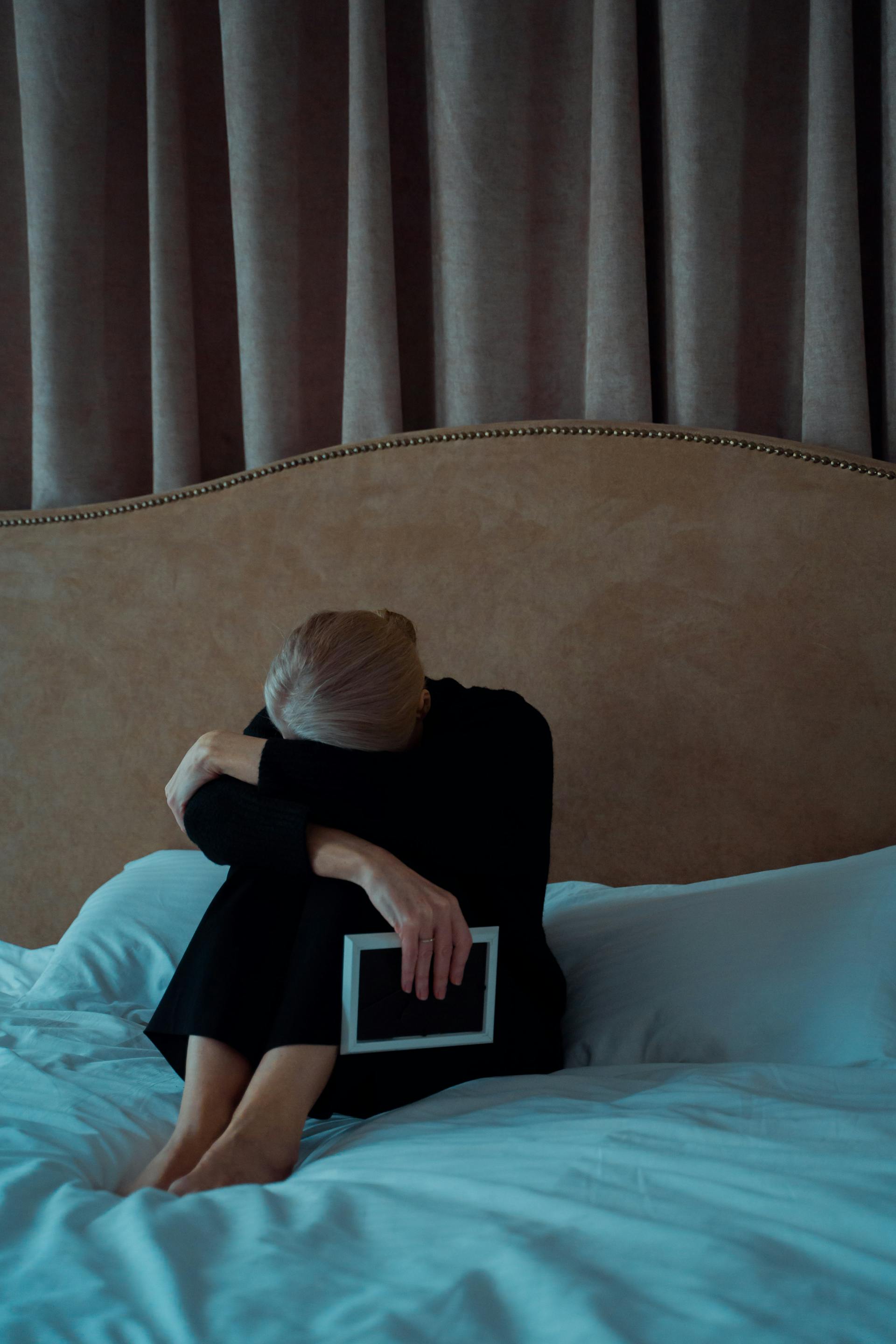 Eine traurige ältere Frau hält einen Fotorahmen, während sie auf ihrem Bett sitzt | Quelle: Pexels