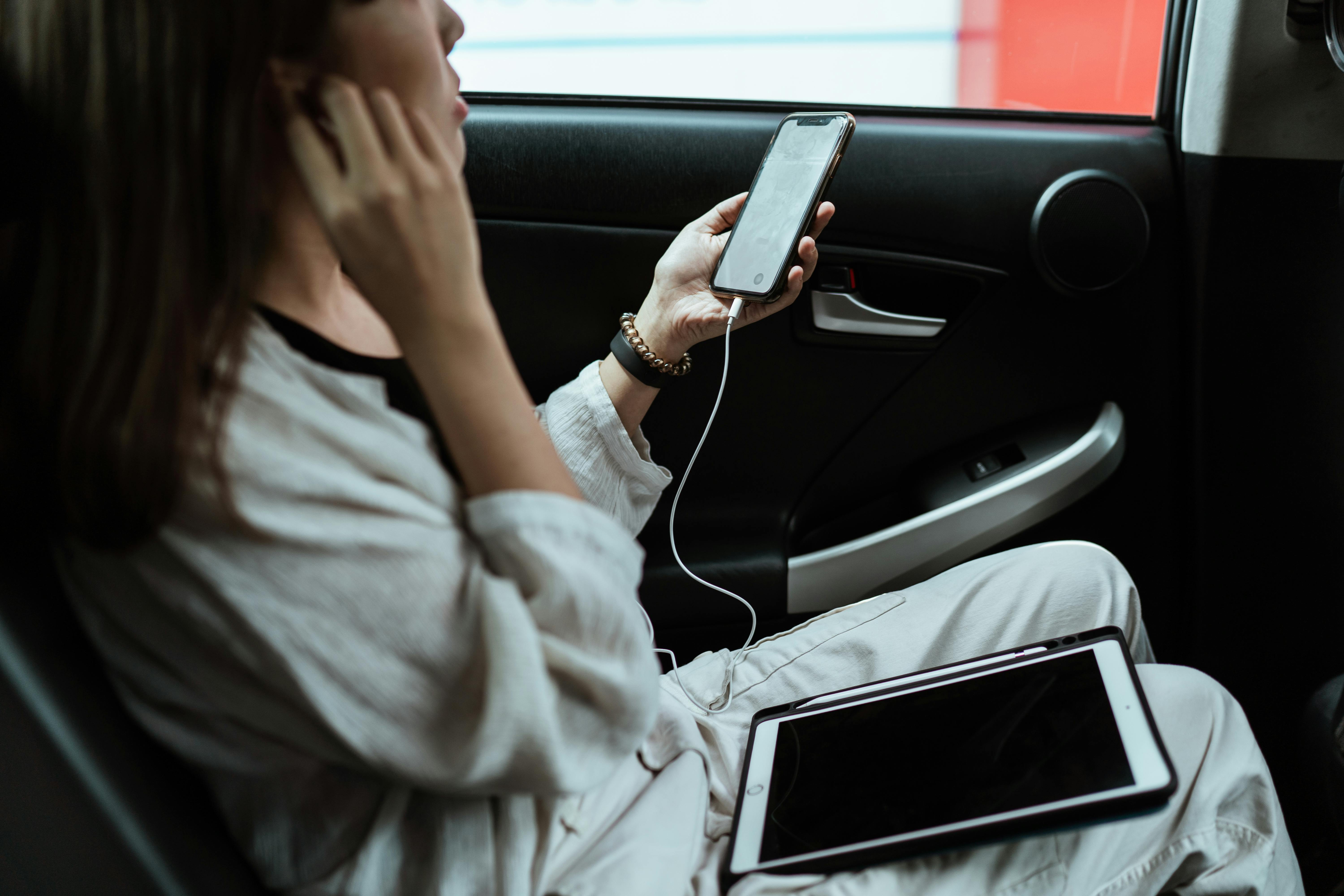 Eine Frau sitzt in einem Auto, hält ein Telefon in der Hand, trägt Kopfhörer und hat einen Laptop auf dem Schoß | Quelle: Pexels