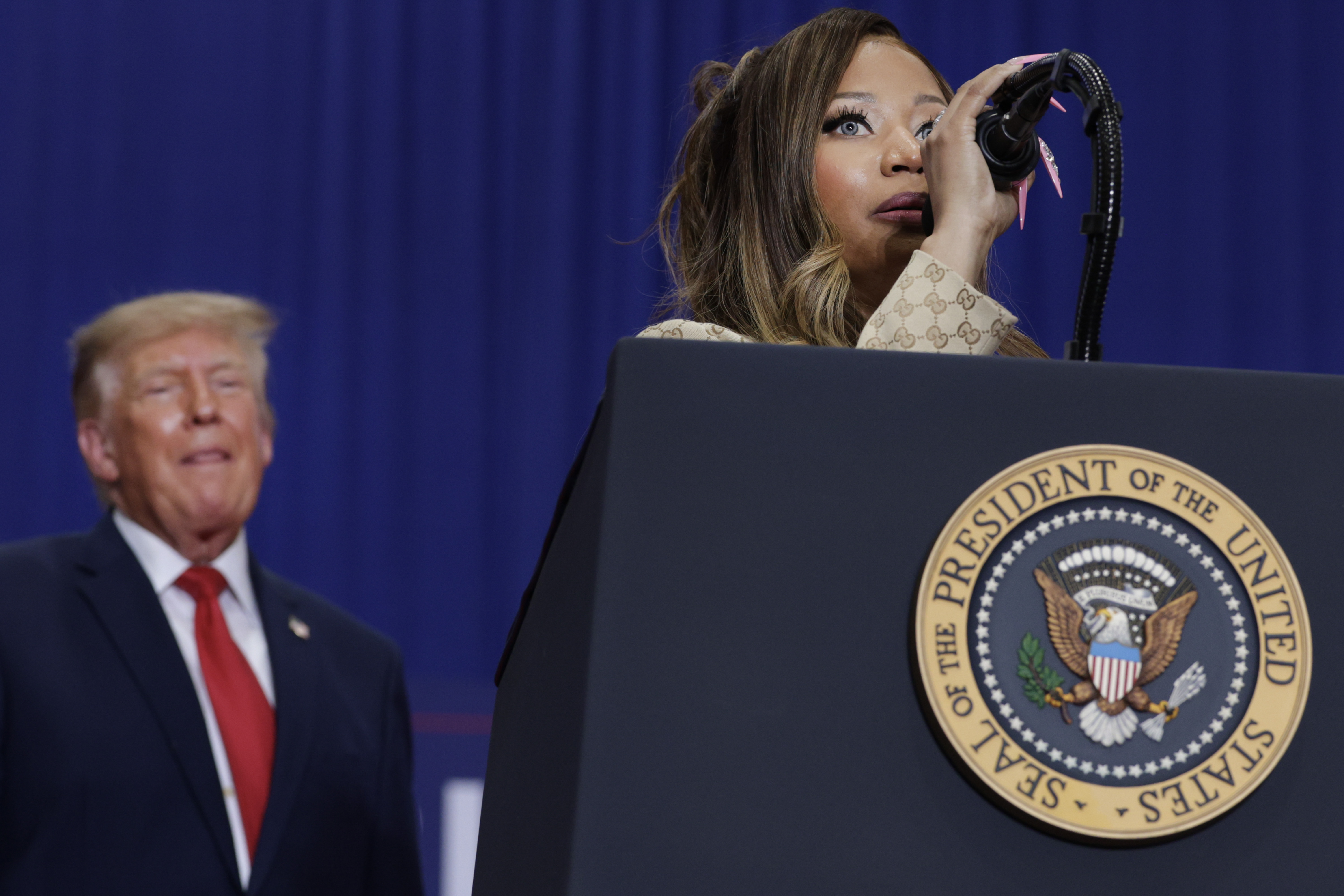 Nicki Minaj spricht hinter einem Rednerpult mit dem Präsidentenwappen, während Donald Trump von der Seite zusieht – ein seltener Moment, in dem die Rapperin bei einer Wahlkampfveranstaltung des Präsidenten statt bei einem Konzert im Mittelpunkt steht. | Quelle: Getty Images
