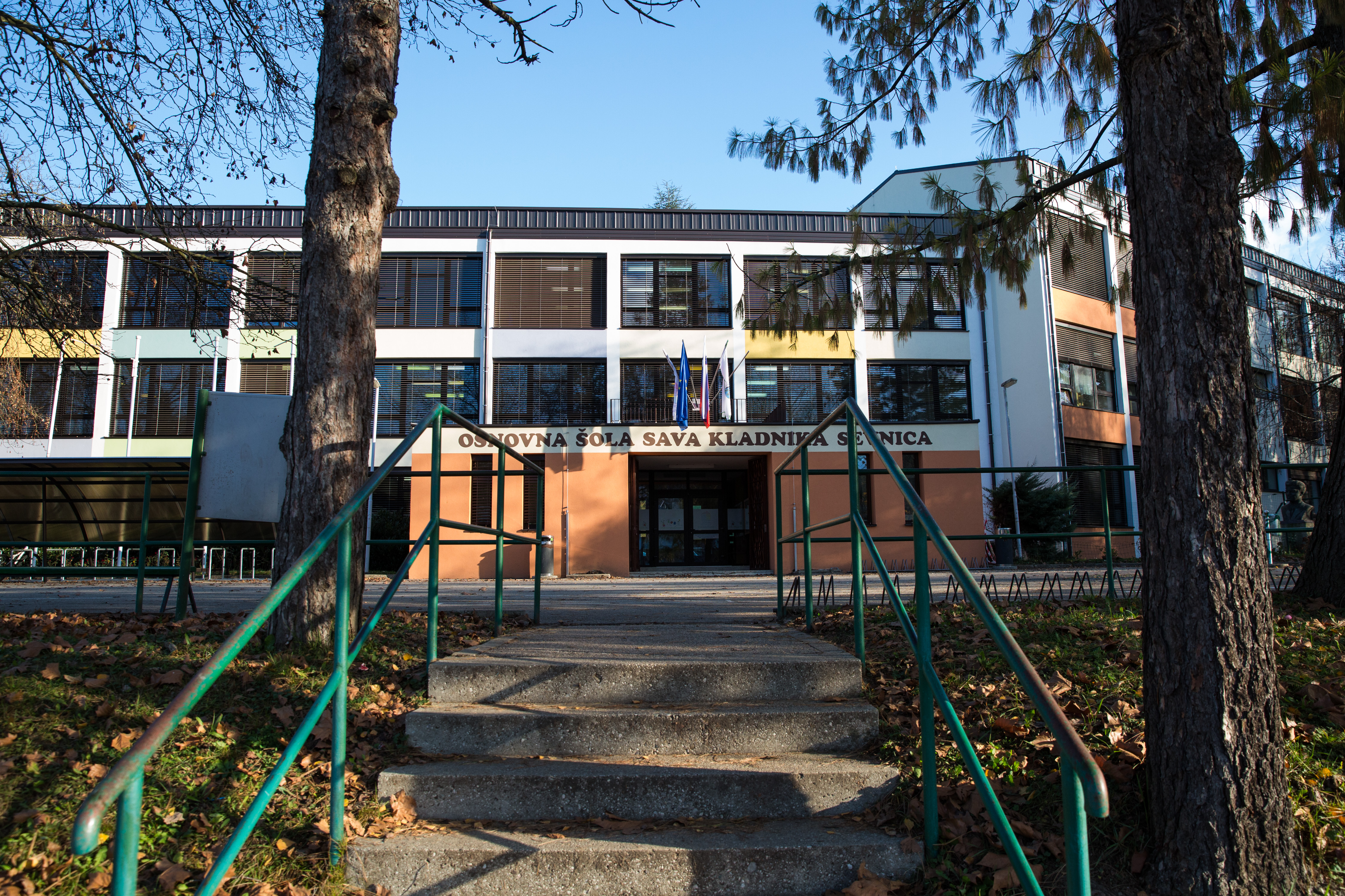 Ein weitläufiger Landschaftsblick, aufgenommen am 29. November 2016, zeigt Betonstufen mit grünen Geländern, die zur „Osnovna šola Sava Kladnika“ hinaufführen – der Grundschule in Sevnica, Slowenien, die Melania Trump besucht hat. Umgeben von hohen immergrünen Bäumen und verstreuten Herbstblättern steht das mehrstöckige Schulgebäude friedlich unter strahlend blauem Himmel. | Quelle: Getty Images