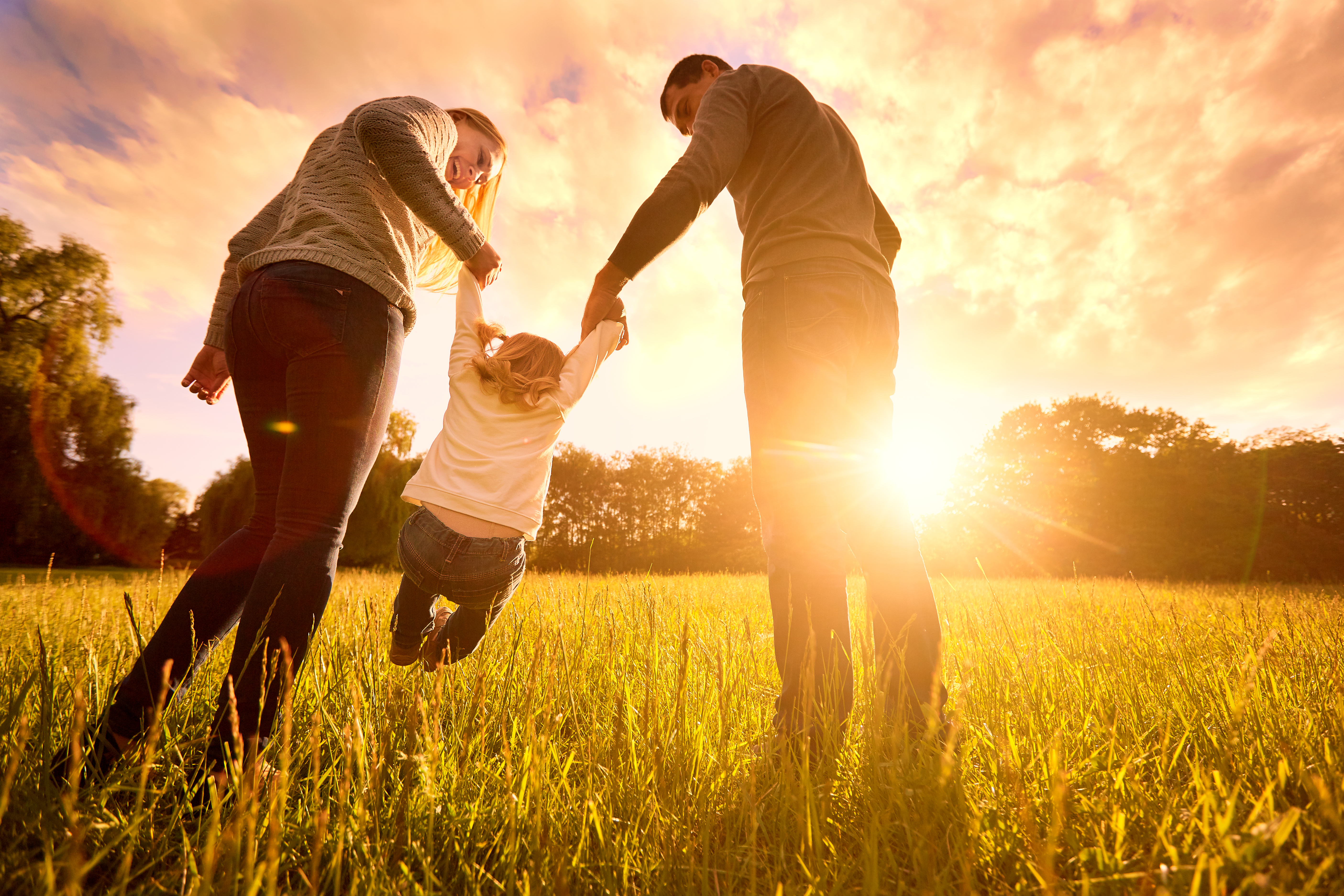 Ein Mann und eine Frau, die auf einer Wiese vor dem Hintergrund des Sonnenuntergangs ein Kind in die Luft heben | Quelle: Shutterstock