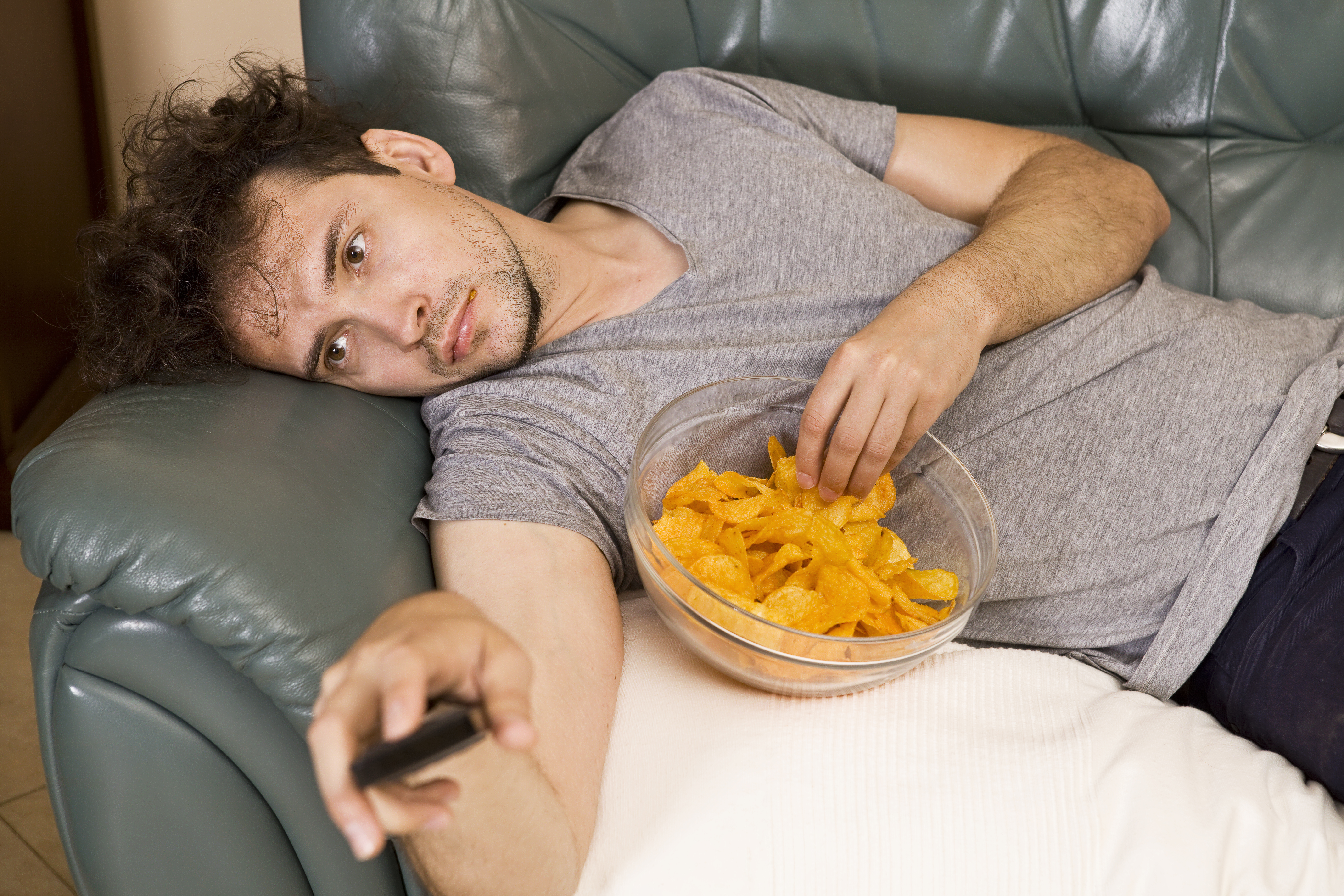 Ein fauler Mann, der auf einer Couch liegt, fernsieht und Snacks isst | Quelle: Getty Images