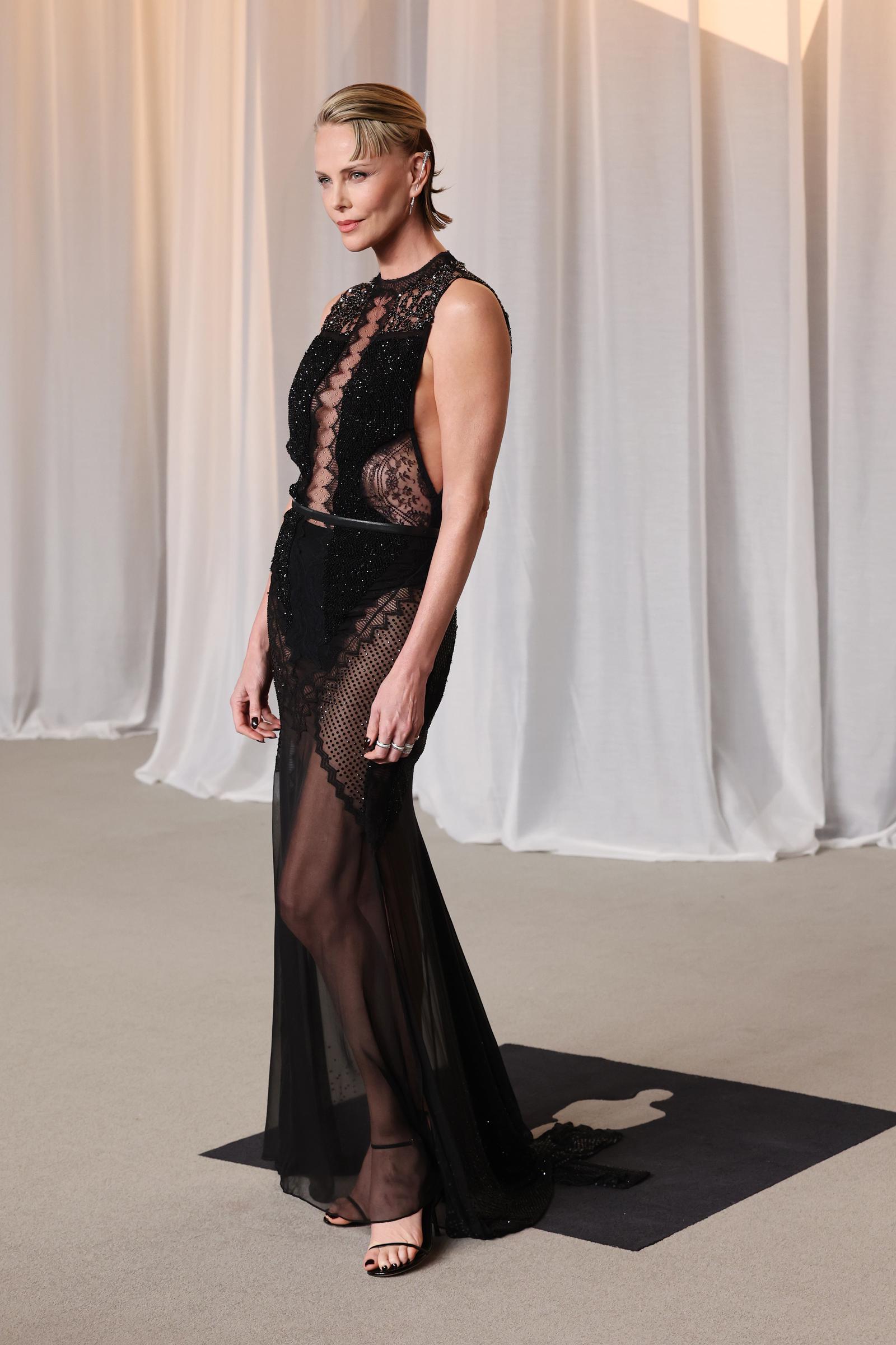Charlize Theron sorgt auf dem roten Teppich für ein kühnes Drama in einem ärmellosen, transparenten schwarzen Abendkleid, das aufwendig mit zarter Spitze, Perlenstickerei und Netz-Einsätzen verziert ist. Betont durch einen schmalen Taillengürtel und kombiniert mit einer glatten Hochsteckfrisur im Wet-Look sowie minimalistischen schwarzen Sandalen, verbindet die Oscar-Preisträgerin kantigen, modernen Glamour mit der klassischen Ausstrahlung eines Filmstars, während sie die Verleihung des Icon Award feiert.