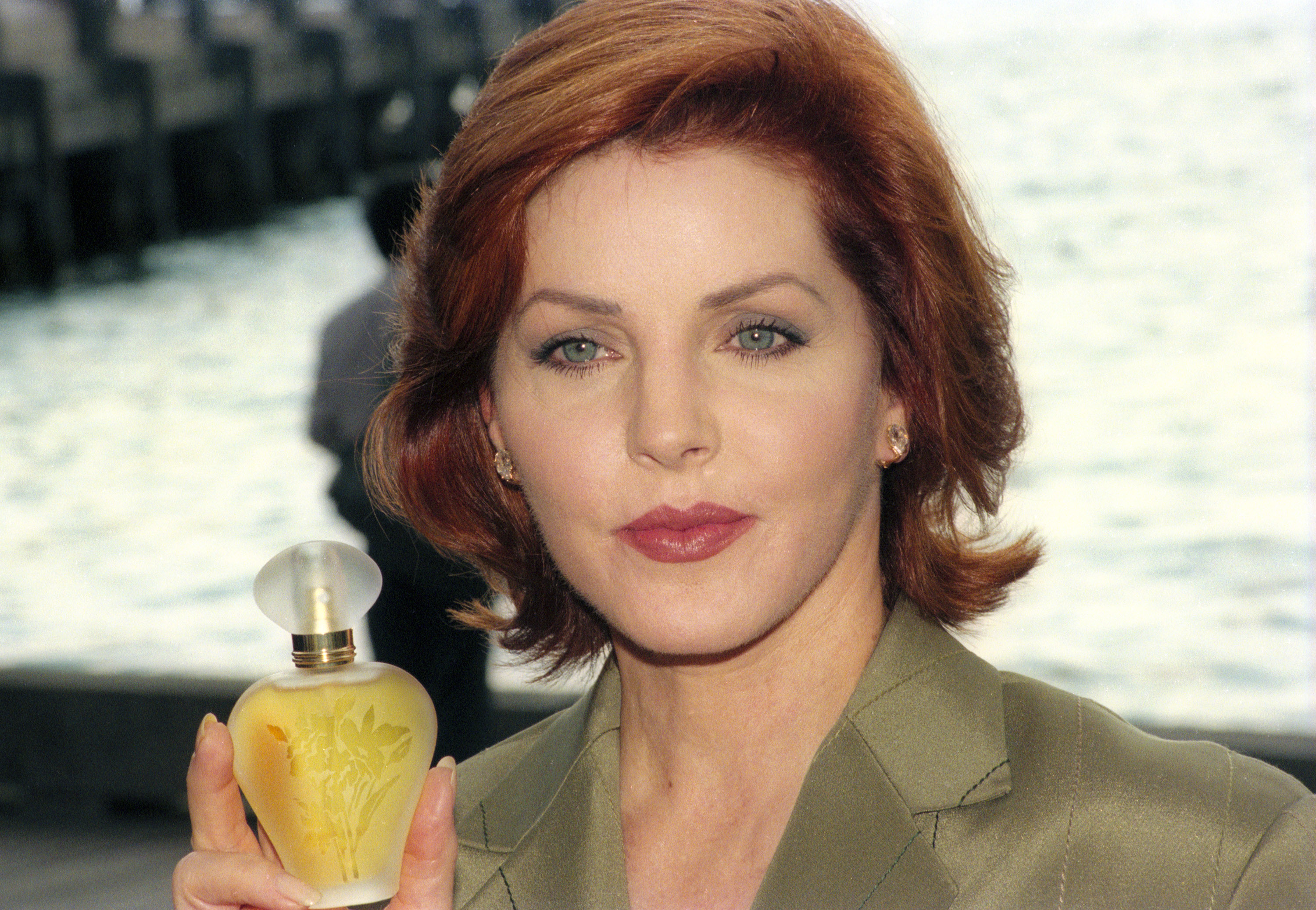 Priscilla Presley hält bei der Vorstellung ihres Parfüms „Moments“ am 29. Oktober 1996 im Park Hyatt Hotel in Sydney, Australien, eine Milchglasflasche in die Höhe. Sie trägt eine olivgrüne, maßgeschneiderte Jacke und hat ihr leuchtend rotbraunes Haar zu einem weichen, nach außen gewellten Bob gestylt. | Quelle: Getty Images