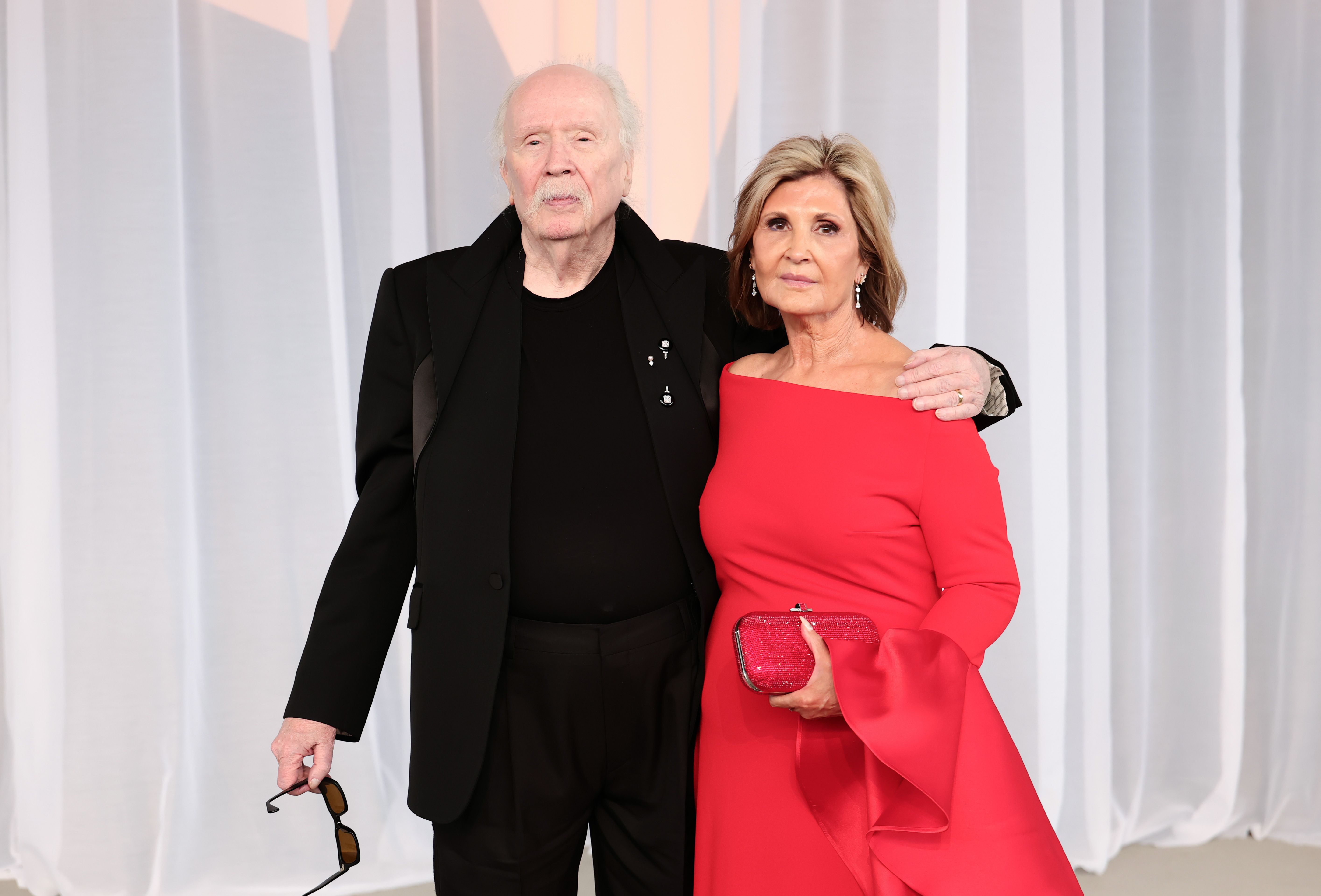 John Carpenter und seine Frau, die Produzentin Sandy King, posieren gemeinsam in aufeinander abgestimmten Outfits. King sticht in einem leuchtend kirschroten, schulterfreien Abendkleid mit dramatischen Glockenärmeln hervor, dazu trägt sie eine funkelnde rubinrote Clutch, während Carpenter sich für einen komplett schwarzen, maßgeschneiderten Anzug mit markanten Revers und einem passenden dunklen Rundhals-Pullover entscheidet. Er hält seine Sonnenbrille an der Seite, während er die Verleihung des Luminary Award feiert.