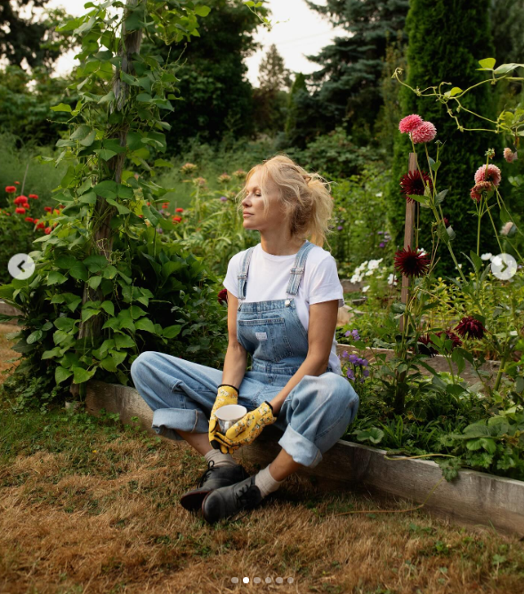 Pamela Anderson hält in ihrem Garten einen Moment inne, um in Ruhe nachzudenken. Sie sitzt mit gekreuzten Beinen auf einem Hochbeet, trägt eine Jeanslatzhose, geblümte Handschuhe und Schnürstiefel und hält eine Tasse in der Nachmittagssonne in den Händen. Umgeben von hohen, blühenden Dahlien, üppigem Grün und Stangenbohnen teilte sie diese friedliche Szene zusammen mit einem Kommentar, in dem sie ihre Kochserie „Pamela’s Cooking with Love“ als „Spaß, Spaß, Spaß“ feierte. | Quelle: Instagram/pamelaanderson
