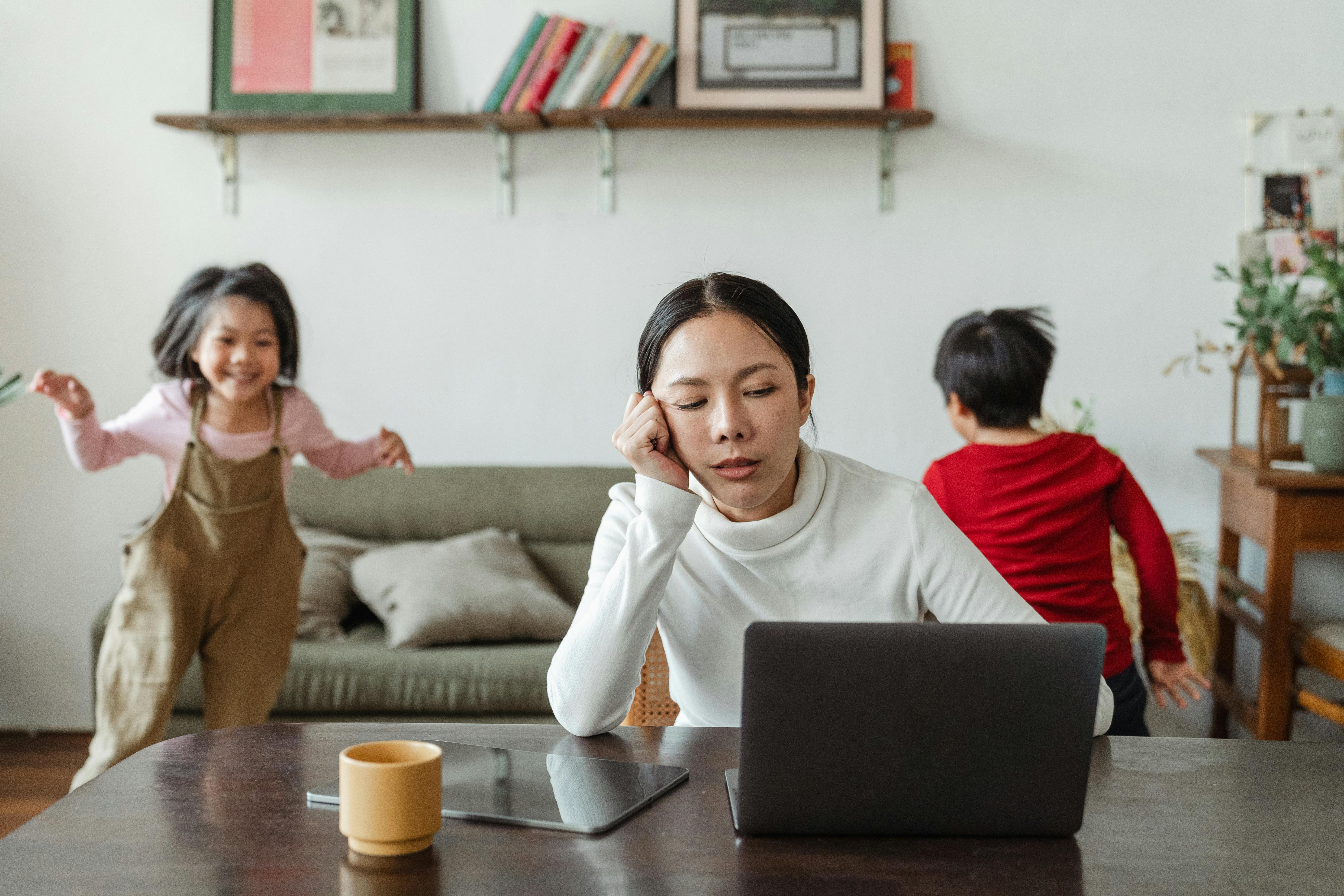 Eine erschöpfte Frau sitzt vor einem Computer, während ihre Kinder im Hintergrund spielen | Quelle: Pexels