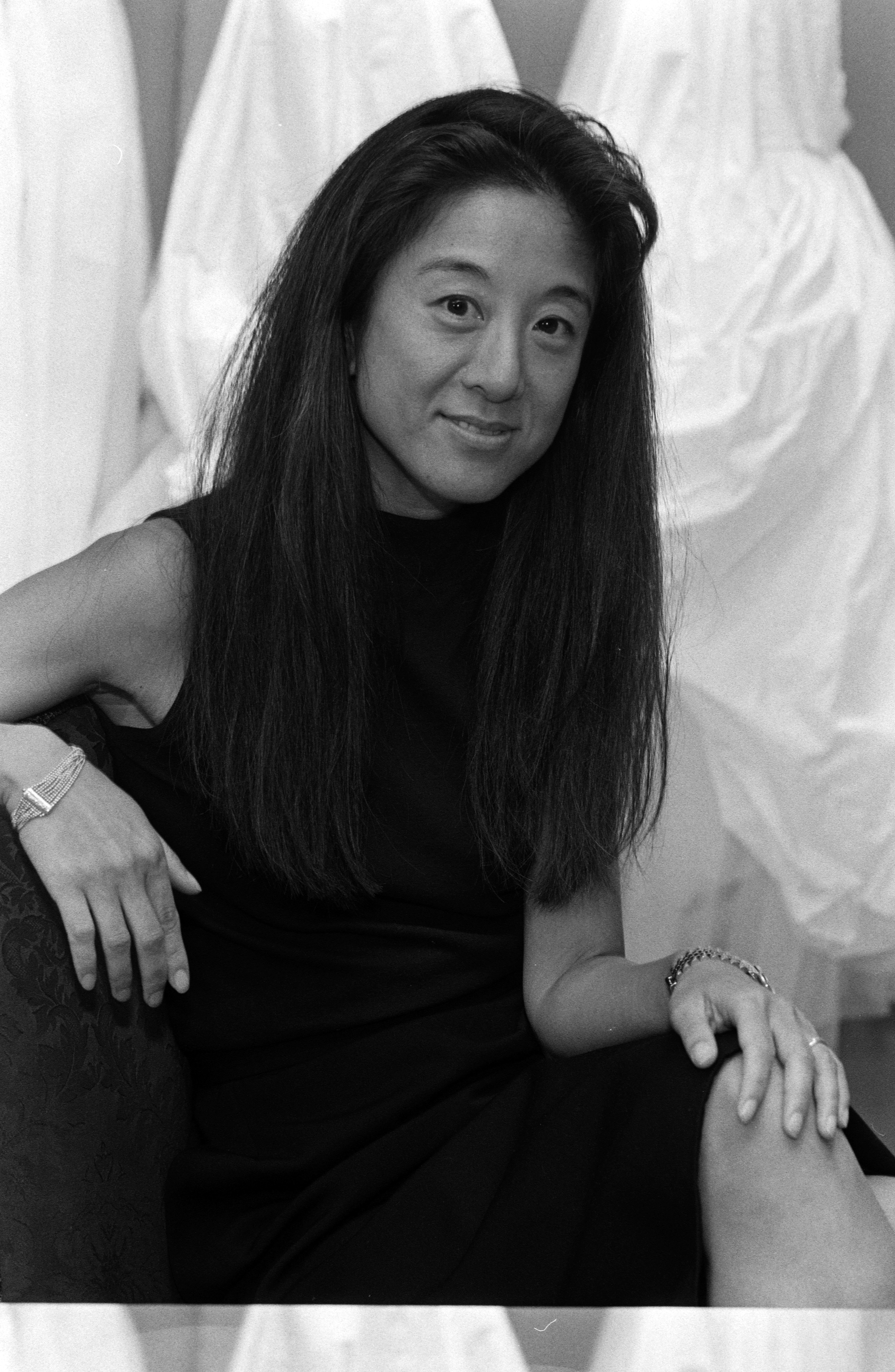 Die Modedesignerin Vera Wang posierte am 22. August 1990 für ein Schwarz-Weiß-Porträt während eines Interviews, in dem sie über ihren Einstieg in die Brautmodenbranche und die Eröffnung des „Vera Wang Bridal House“ sprach. Vor drapierten Brautkleidern sitzend lächelte Wang sanft in einem ärmellosen dunklen Kleid, ergänzt durch Armbänder an beiden Handgelenken und ihr charakteristisches langes dunkles Haar, das glatt über ihre Schultern fiel. | Quelle: Getty Images