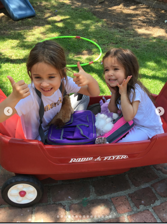Mabel und Fiona genießen einen verspielten Moment in einem großen roten Radio-Flyer-Wagen: Mabel streckt beide Daumen hoch, während Fiona neben ihr lächelt. Zwischen ihnen liegen eine lila Tasche und ein paar Spielsachen, und im sonnigen Garten hinter ihnen sind ein grüner Hula-Hoop-Reifen und eine Kinderrutsche zu sehen. | Quelle: Instagram/mabellowe_