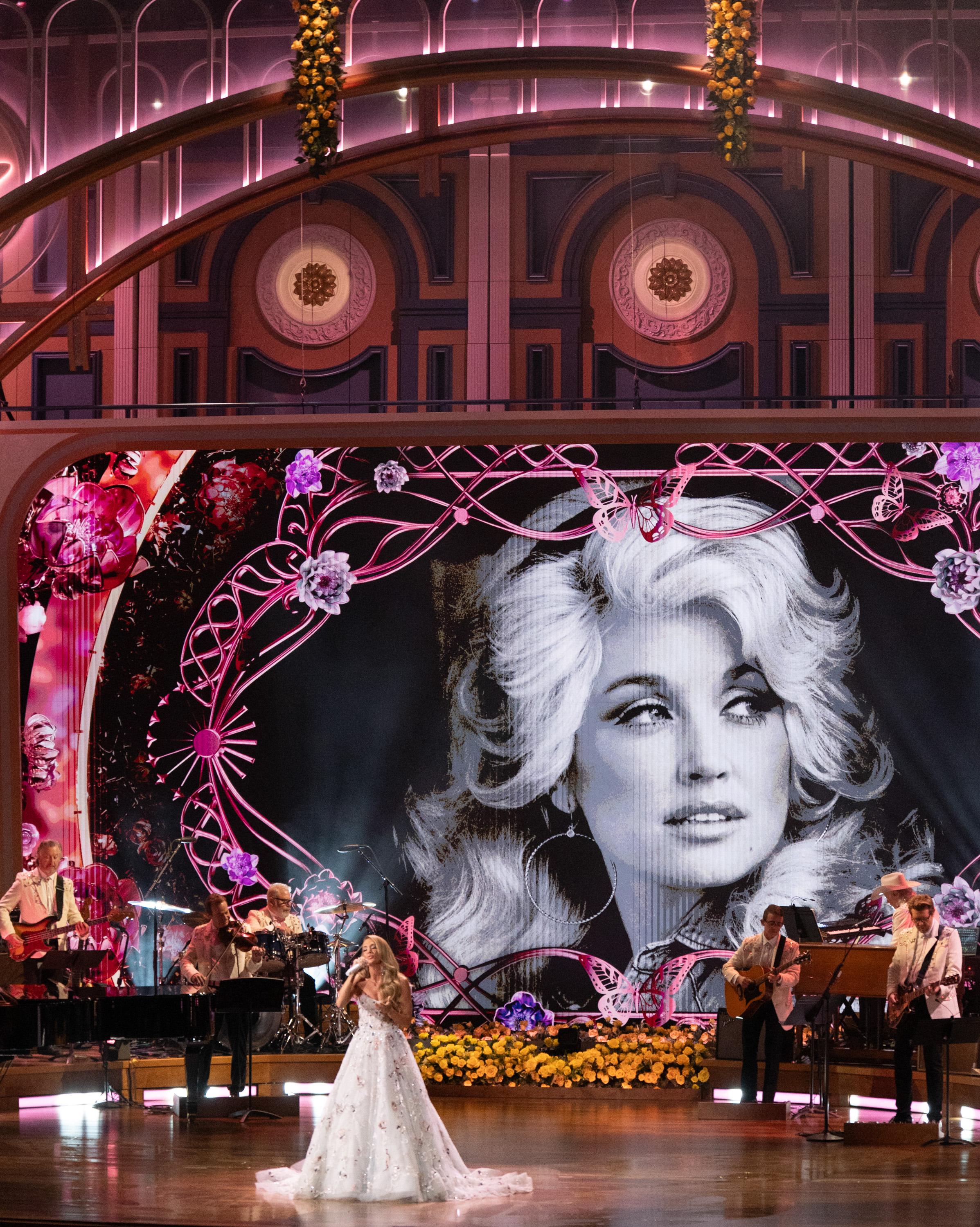 Carrie Underwood tritt während der Hommage in Nashville unter einem riesigen Schwarz-Weiß-Porträt von Dolly Parton auf, umgeben von rosa Schmetterlingen und Blumendekorationen. | Quelle: Getty Images.