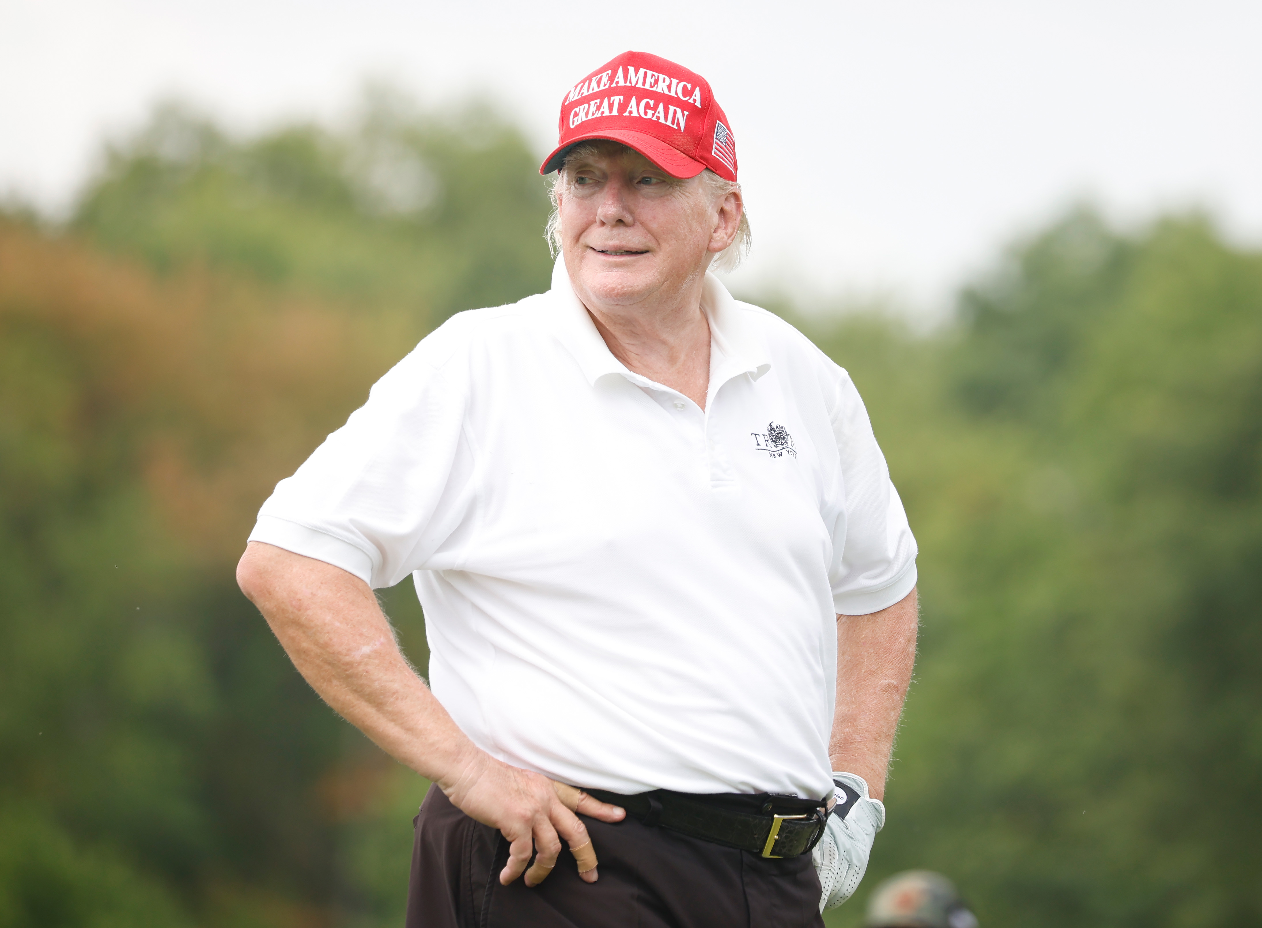 Donald Trump trägt seine charakteristische rote Mütze und sein weißes Polo und steht mit den Händen in den Hüften auf dem Golfplatz. Seine Wangen und seine Kieferpartie sind auffallend rot und kontrastieren mit der blassen Haut an seinem Hals.