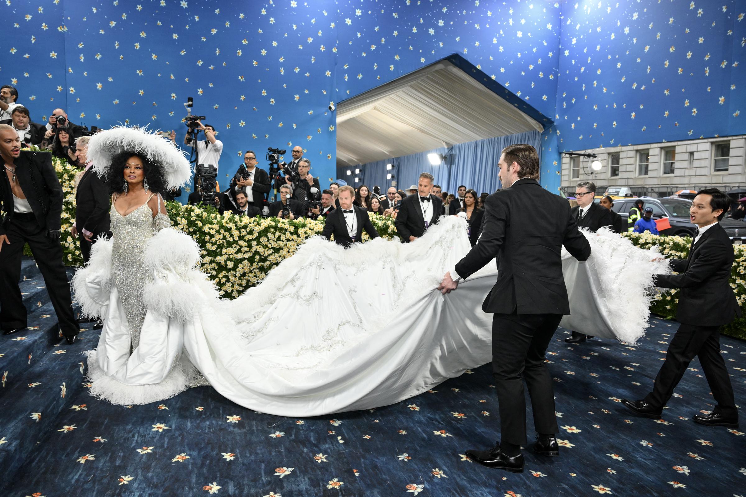 In diesem Moment steht Ross am Fuße der Treppe, während ein Team von Assistenten ihre voluminöse Schleppe vorsichtig anhebt und arrangiert. Das Kleid fächert sich um sie herum auf, seine Stickereien und zarten Strukturen fangen das Licht ein.