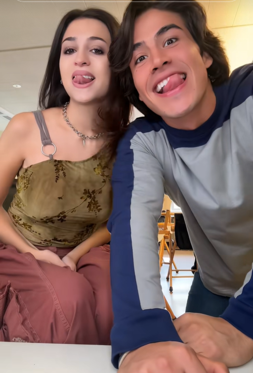 Josie Totah und ihr männlicher Begleiter schneiden in einer weiteren Szene aus dem TikTok-Video verspielte Grimassen in die Kamera. Josie trägt dasselbe olivgrüne Oberteil mit Blumenmuster und dieselbe rosa Hose, dazu eine silberne Halskette und einen Schulterriemen. | Quelle: TikTok/josie totah