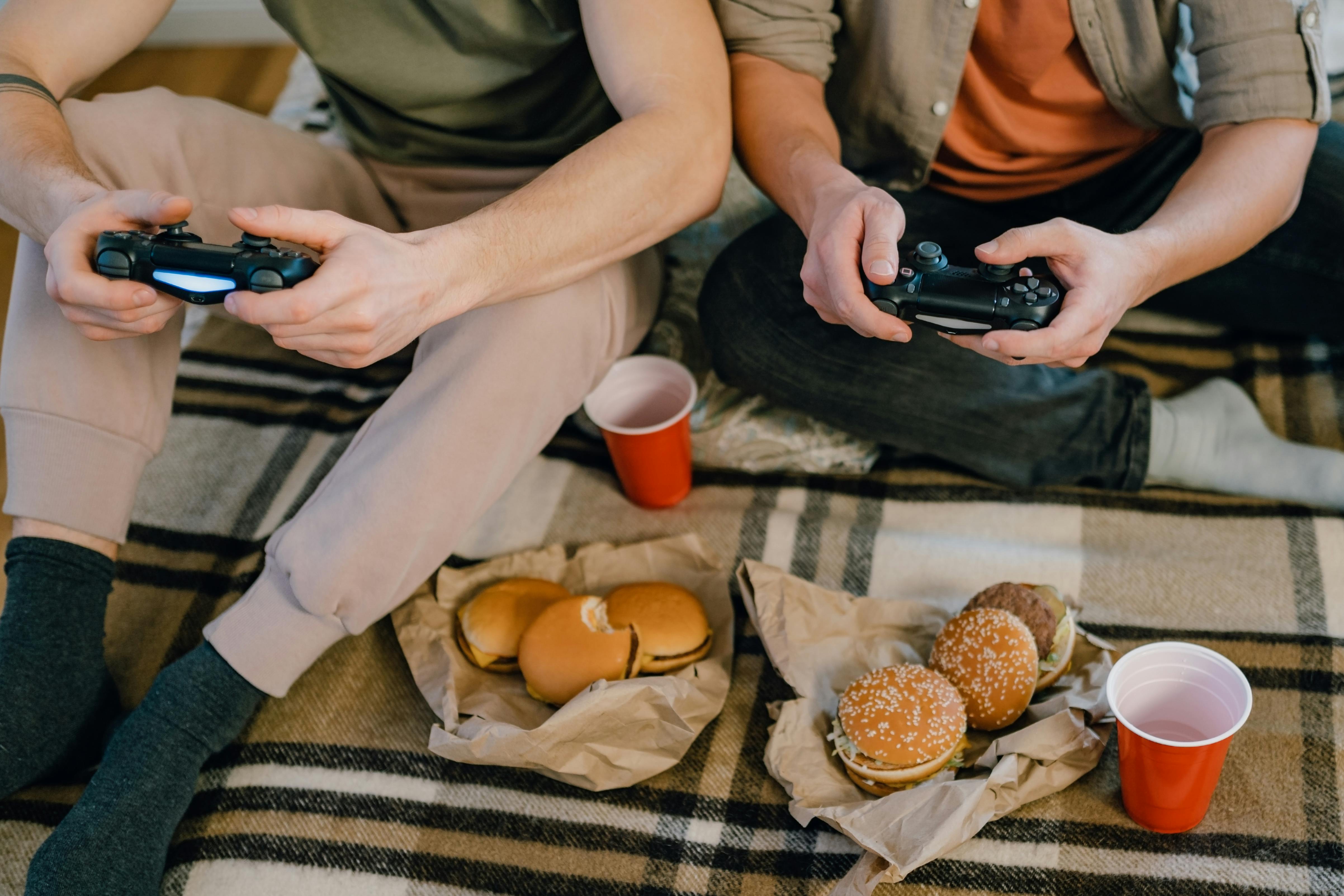 Zwei Männer sitzen auf dem Boden und essen Burger, während sie Videospiele spielen | Quelle: Pexels