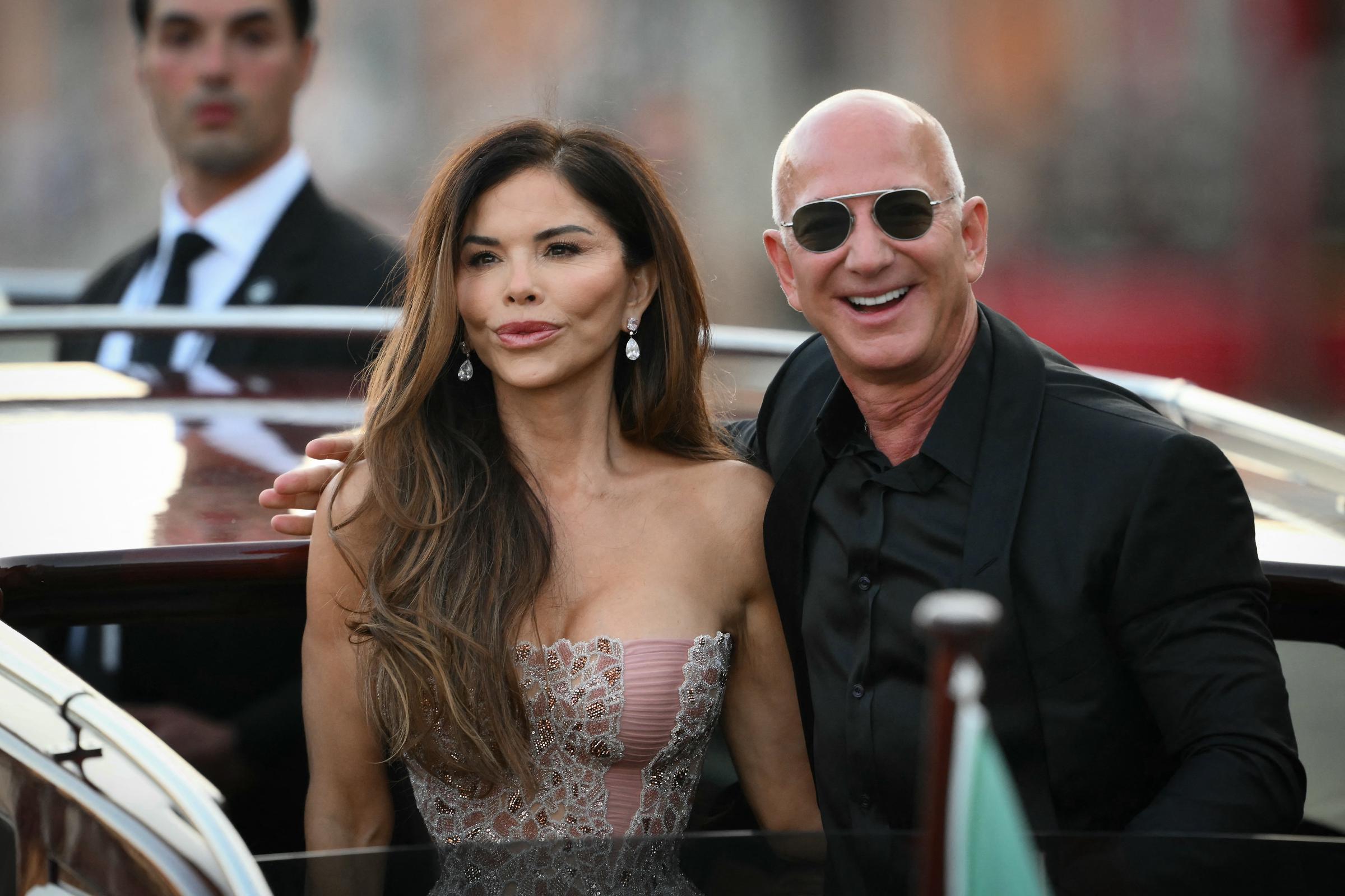 Jeff Bezos zeigt ein breites Lächeln, während er sich dicht an Lauren Sánchez lehnt, die mit ruhiger Miene nach vorne blickt. Das Paar steht auf dem Deck des Bootes und strahlt Zuversicht aus, während ein Sicherheitsbeamter im Anzug von hinten auf die verschwommene Stadtkulisse schaut.